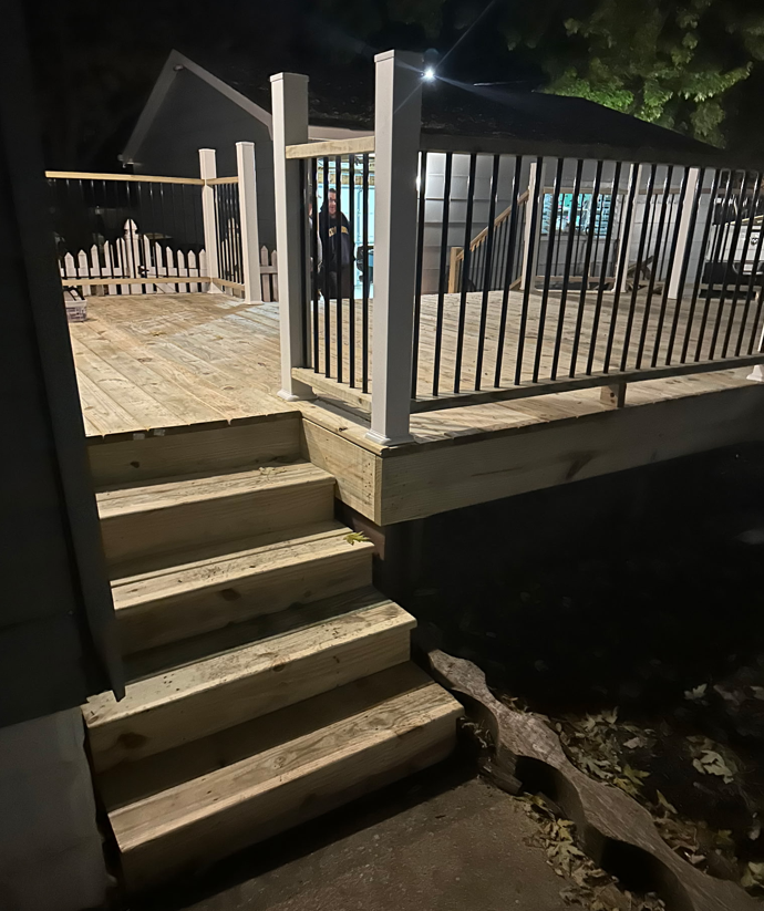 Wooden deck with stairs, black railing, and white posts at night.