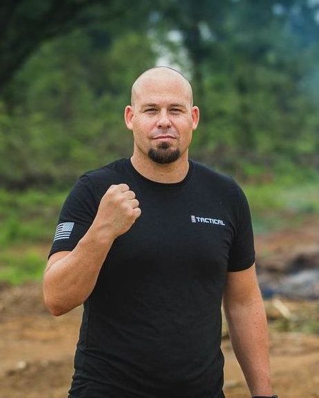 A bald man with a beard wearing a black shirt is standing in a field with his fist in the air.