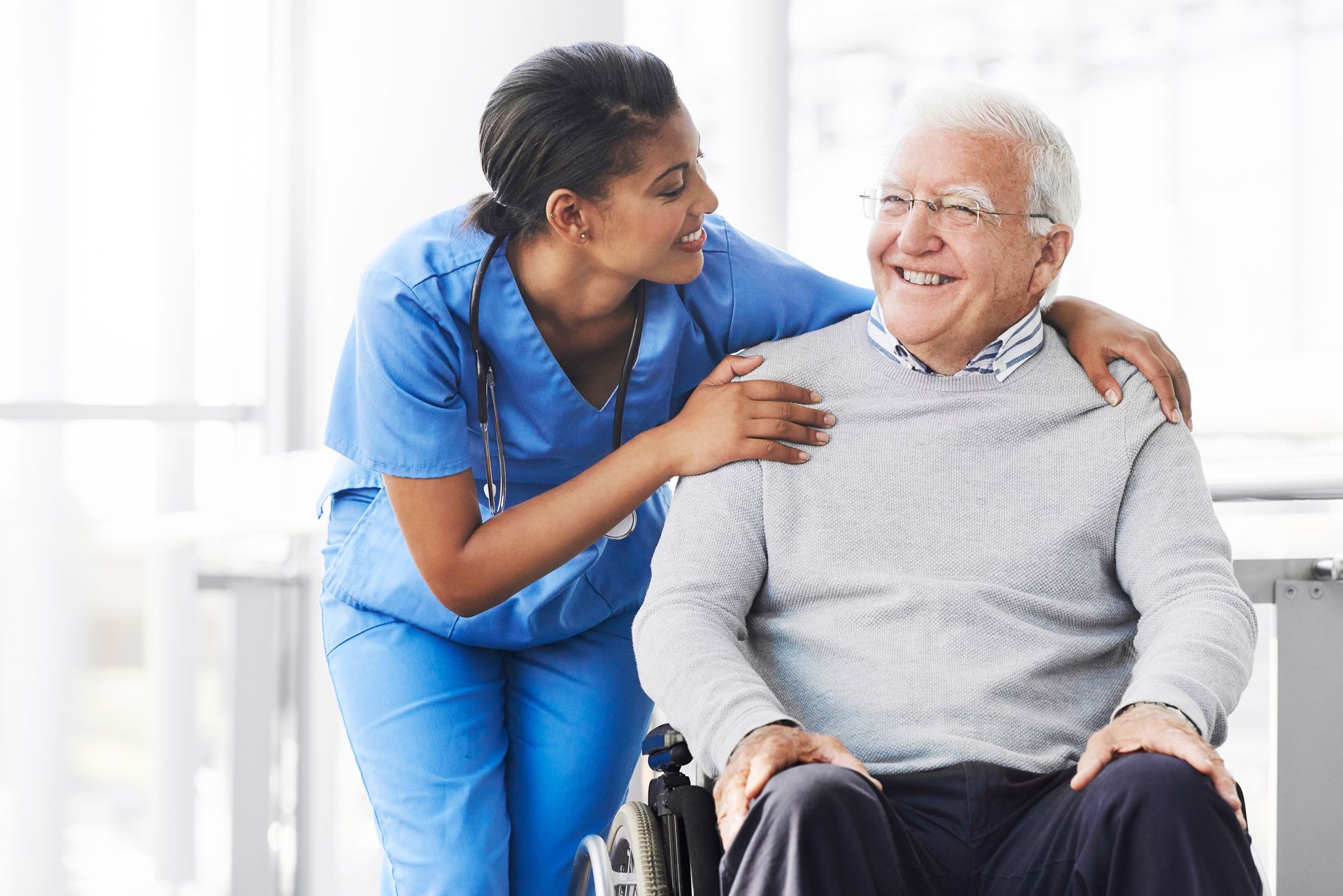 Healthcare worker in scrubs comforting an older adult seated in a wheelchair.