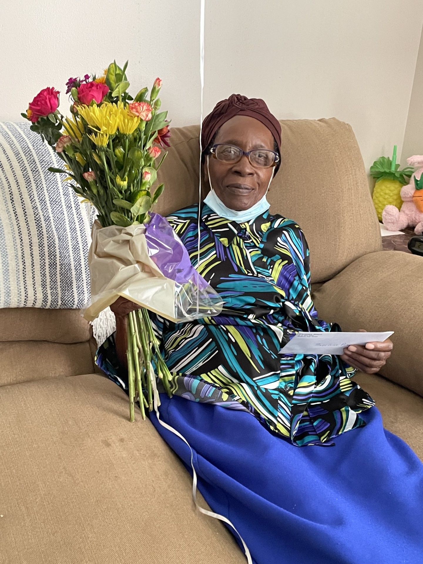 Woman with bouquet and card seated on a couch, wearing patterned shirt and blue skirt.