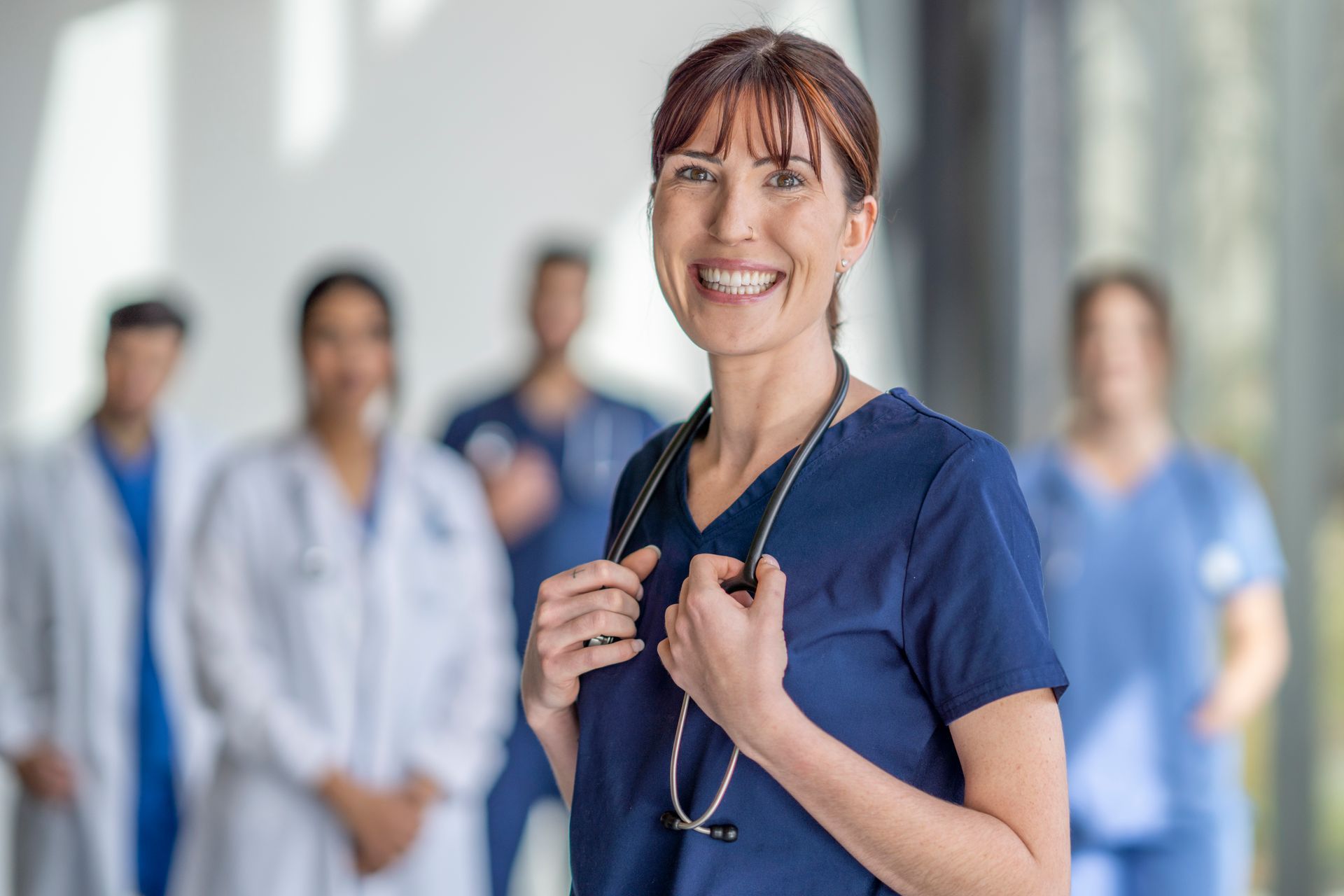 Smiling nurse in blue scrubs holding a stethoscope, with medical staff blurred in the background.