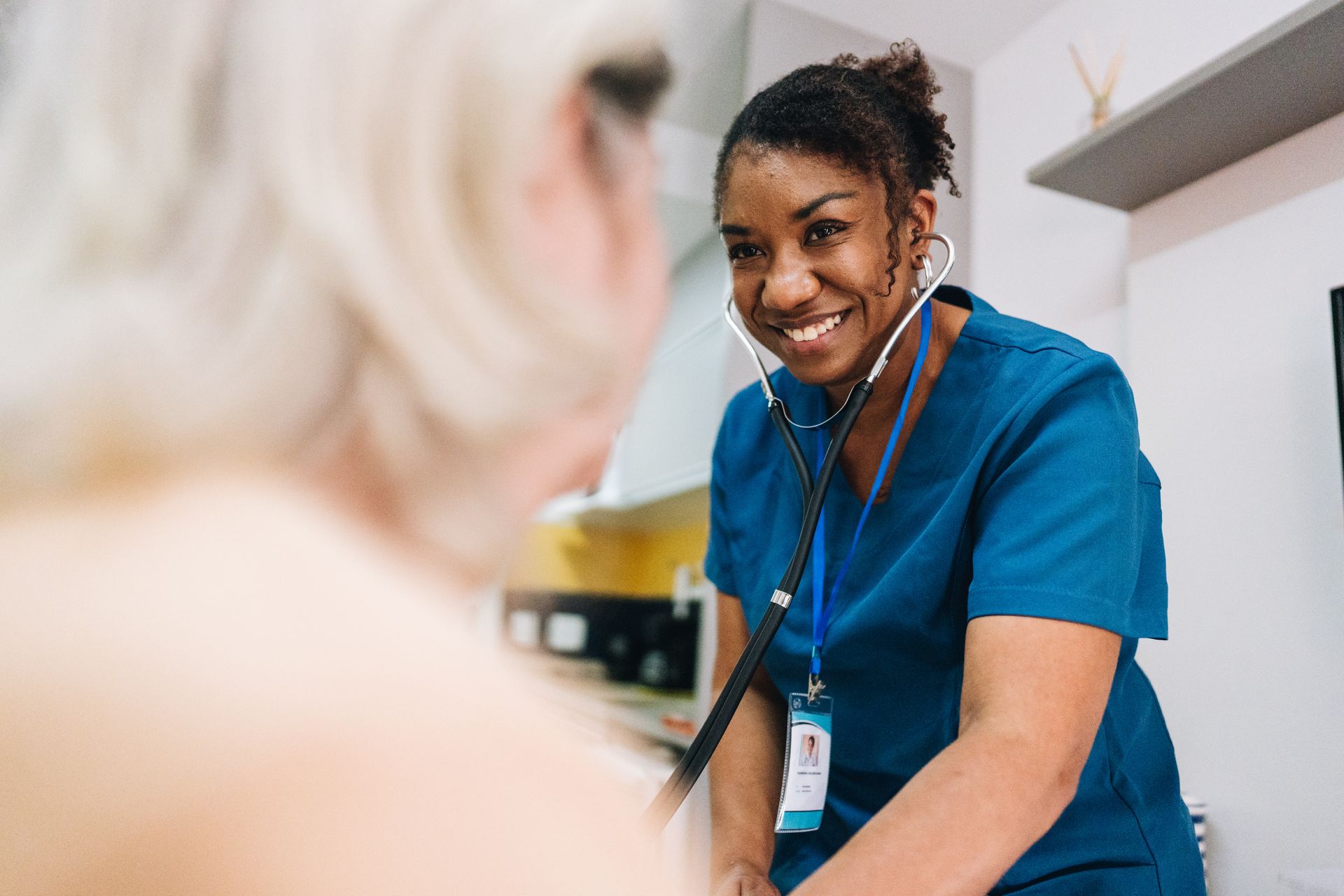 Nurse in blue scrubs smiling while checking a patient with a stethoscope in a clinic room