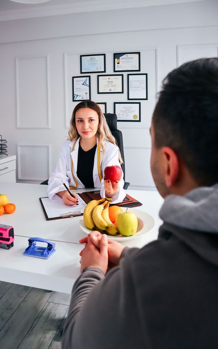 Woman at a table with a bowl of fruit, speaking to a person across from her in an office setting