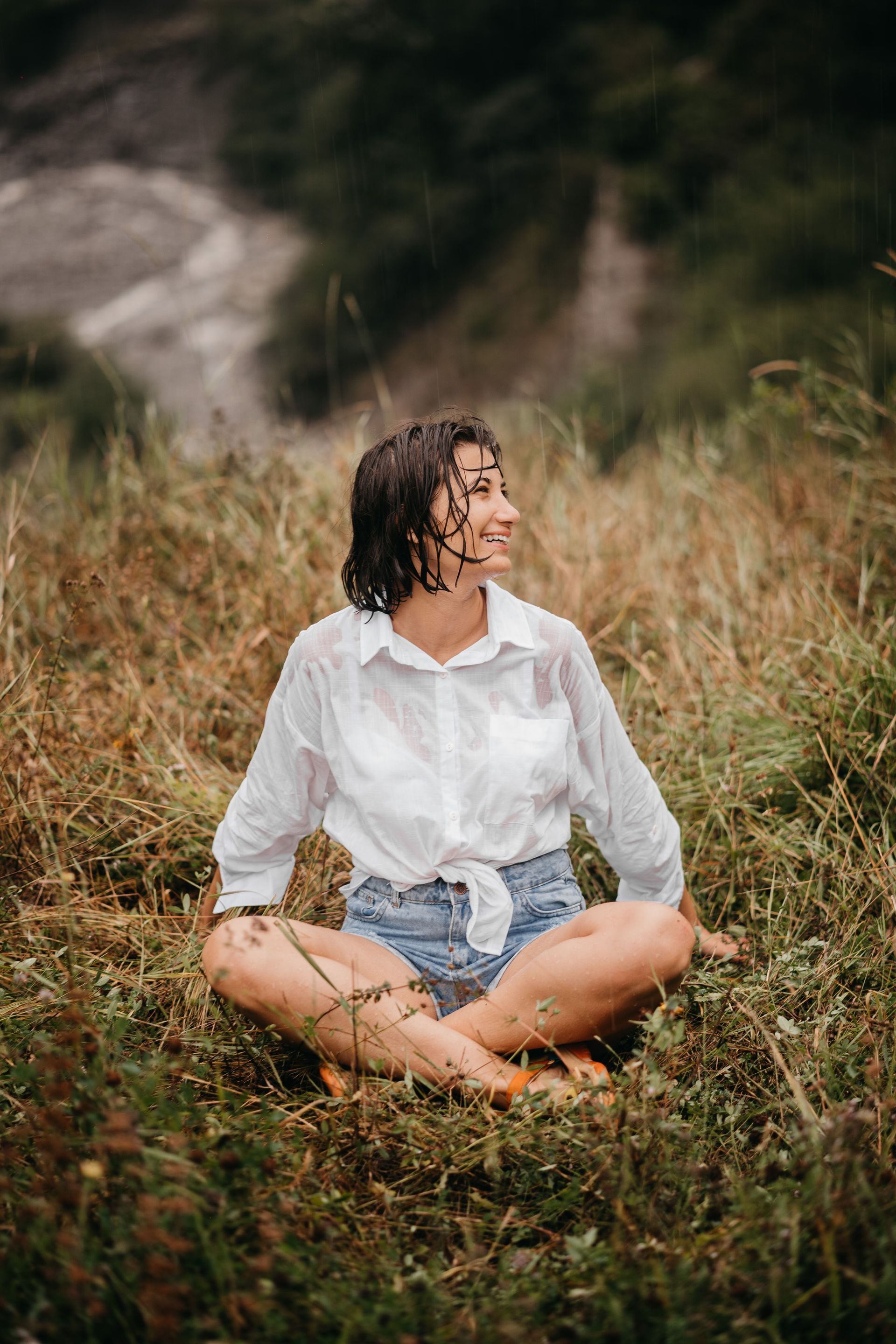 Person in a white shirt and denim shorts sitting cross-legged in tall grass, looking to the side.