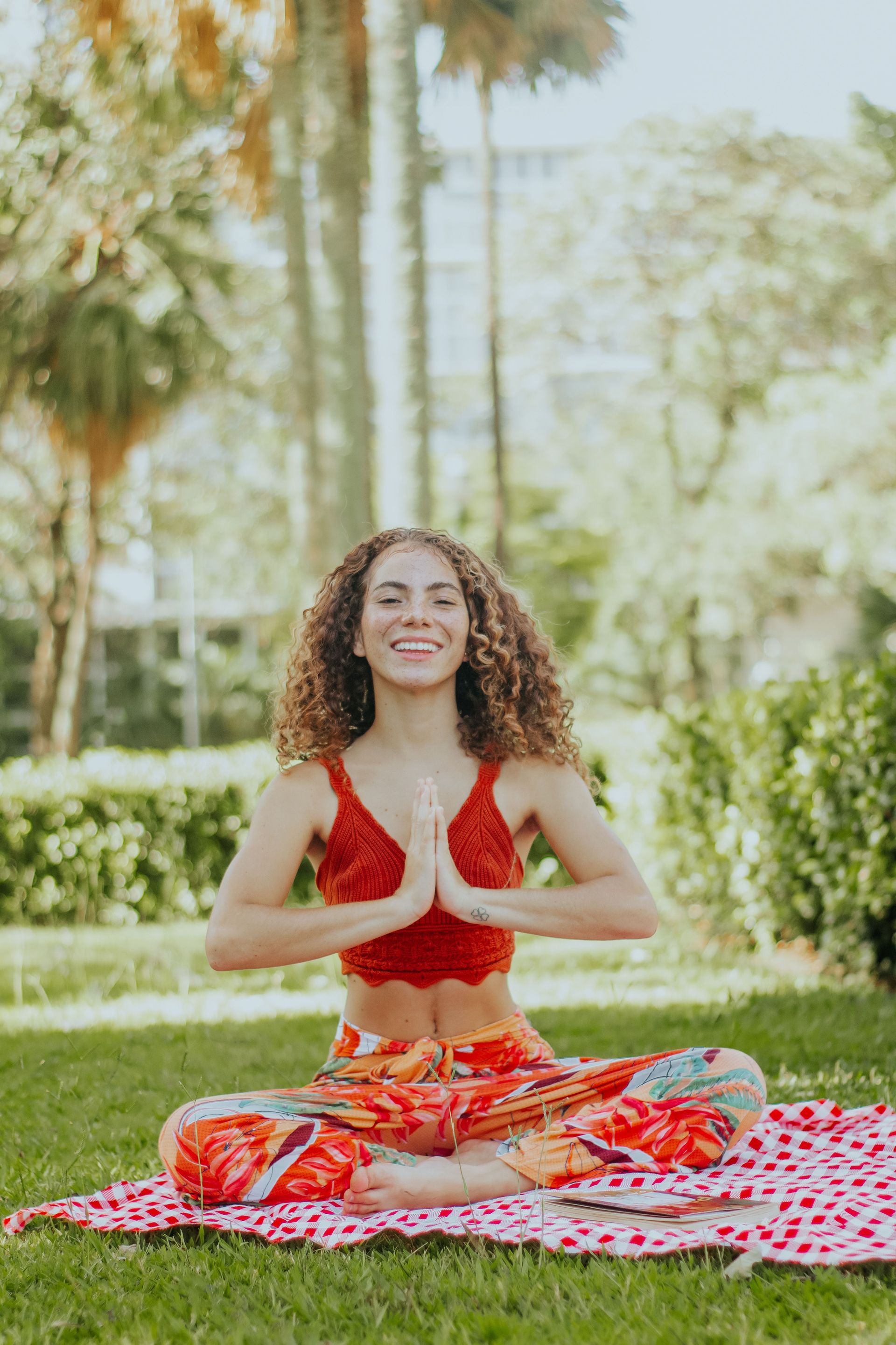Woman meditating cross-legged on a pink blanket in a sunny park, hands in prayer pose.