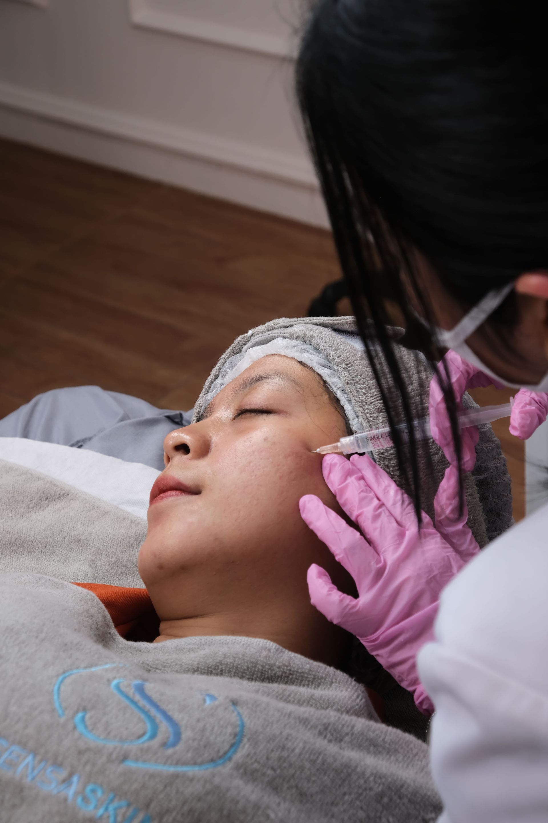 Cosmetician wearing pink gloves applies a facial treatment to a reclining client.