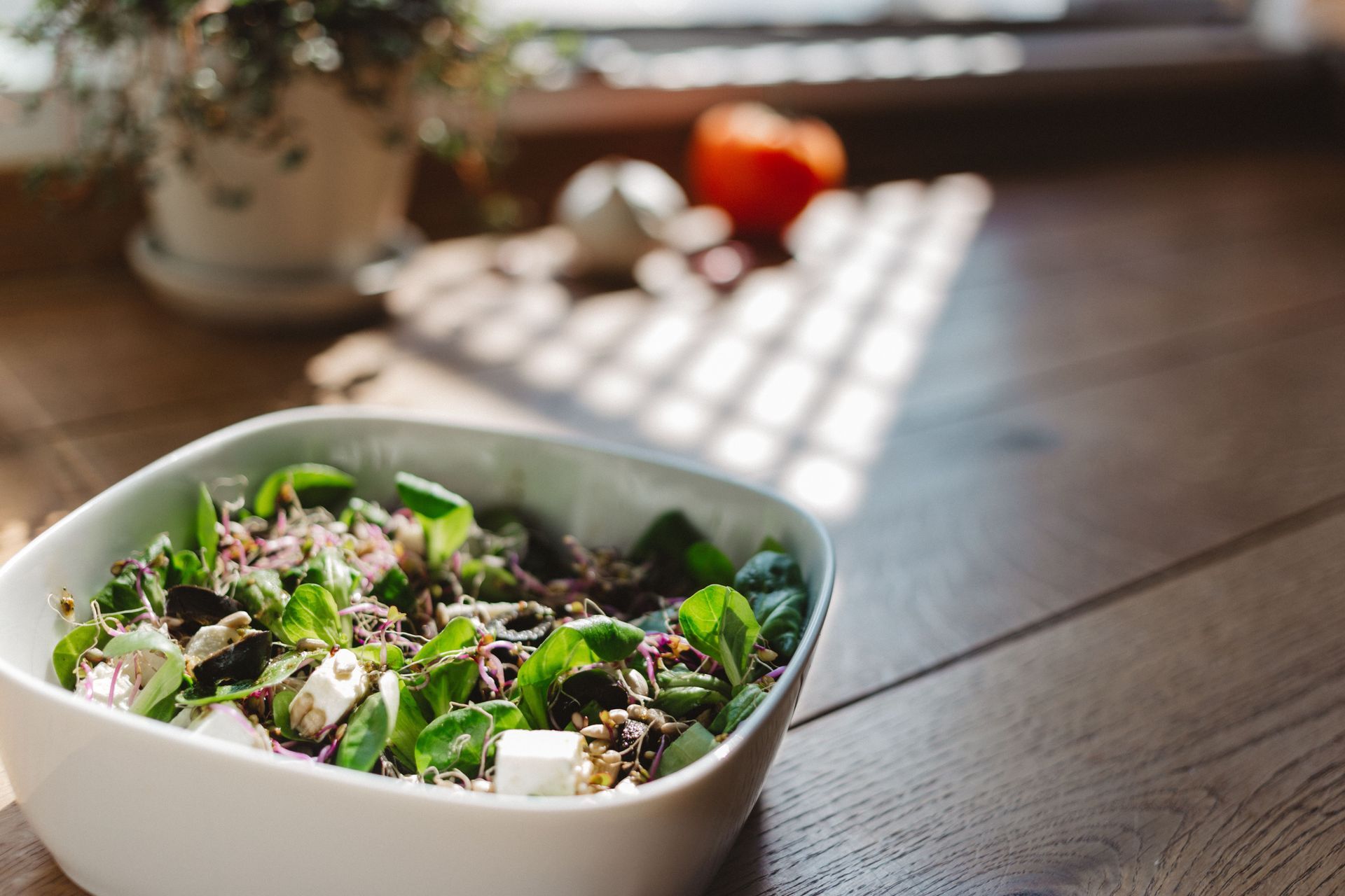 Bowl of fresh salad on a wooden table by a sunny window, with a blurred plant in the background