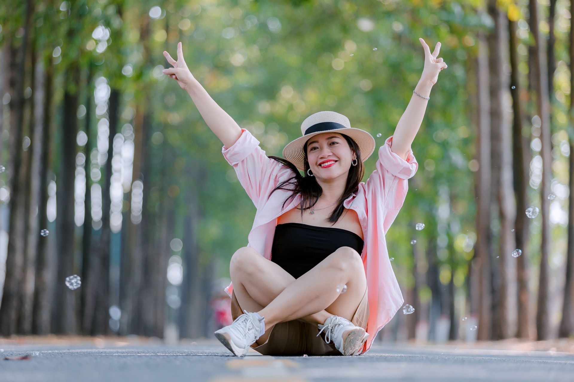 Smiling woman in a hat sits on a path with arms raised, surrounded by tall trees.