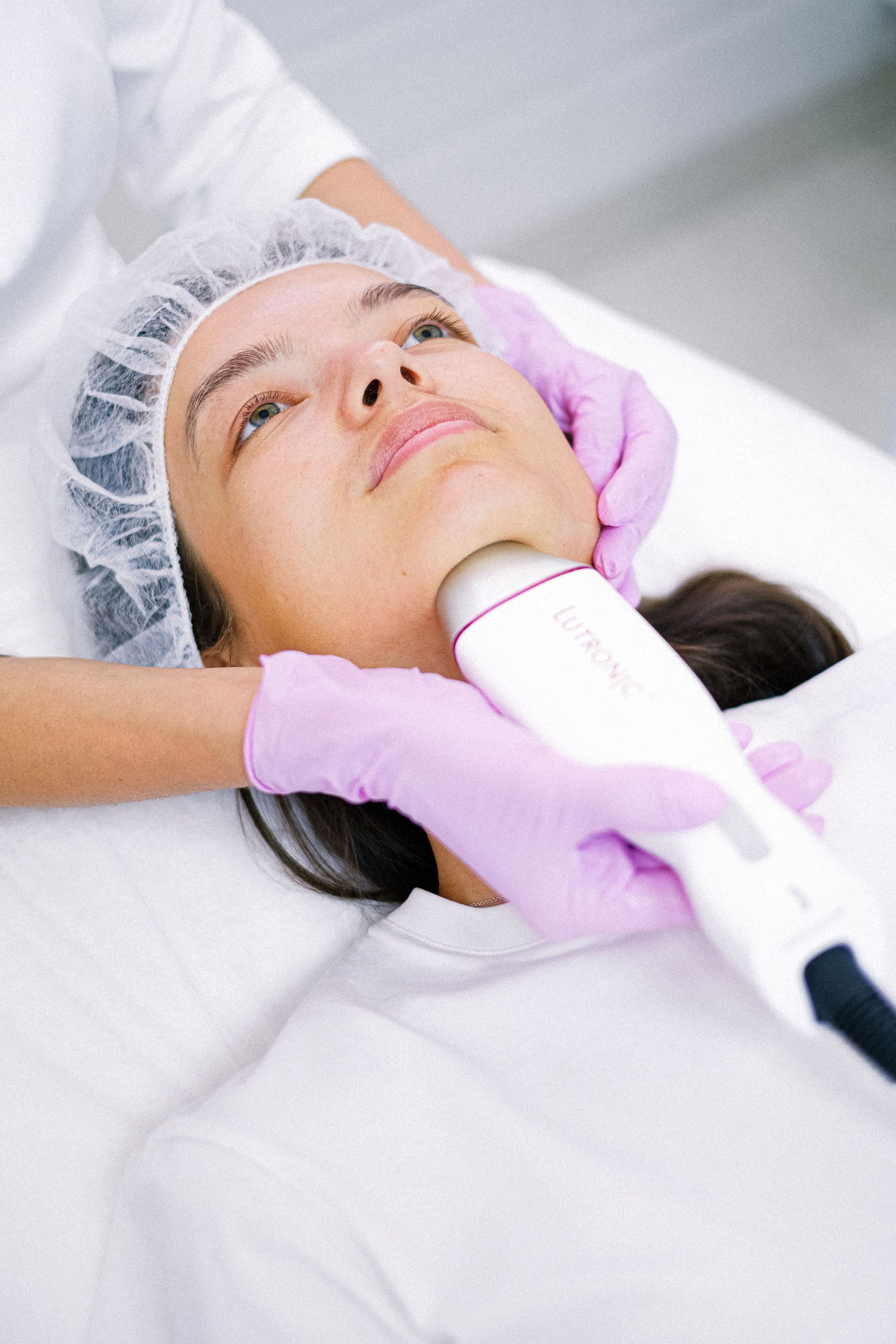A person receiving a facial treatment with a handheld device while lying on a bed, wearing a hair cap and purple gloves.