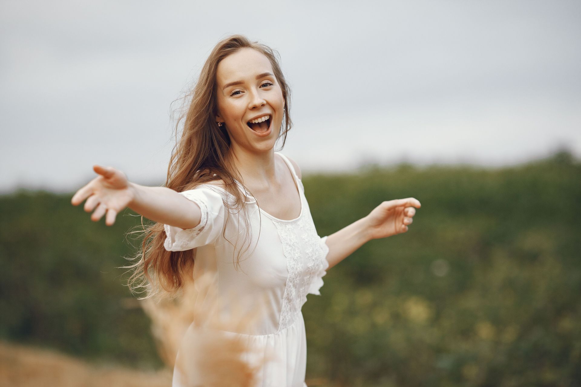 Smiling person in a white dress with arms outstretched in a grassy field outdoors