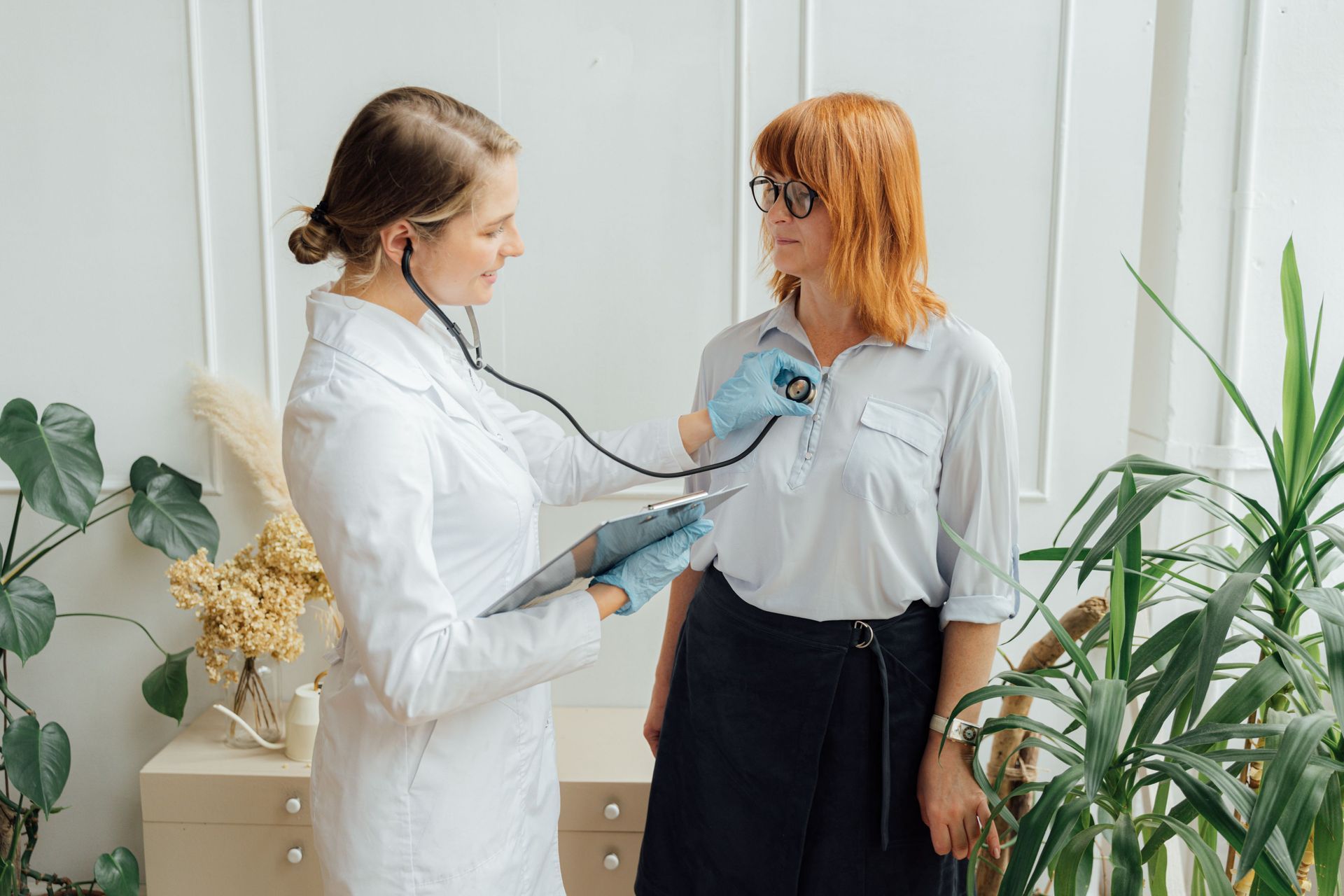 Doctor using a stethoscope to examine a seated woman in a white room