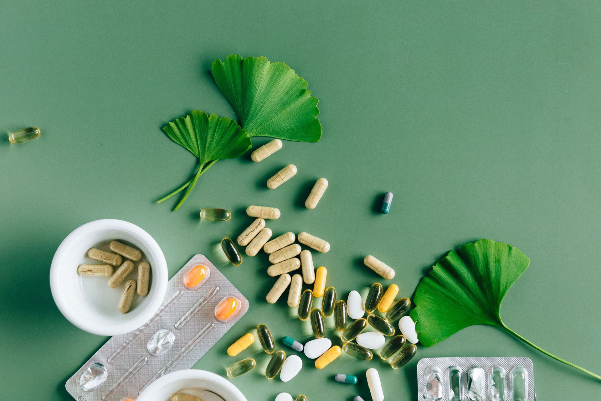 Assorted capsules and tablets spilled on a green background with leaves and a pill tray