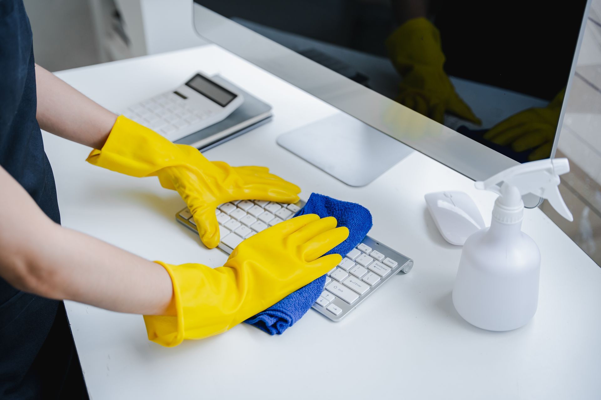Person wearing yellow gloves cleans a keyboard with a blue cloth at a white desk next to a spray bottle and calculator.