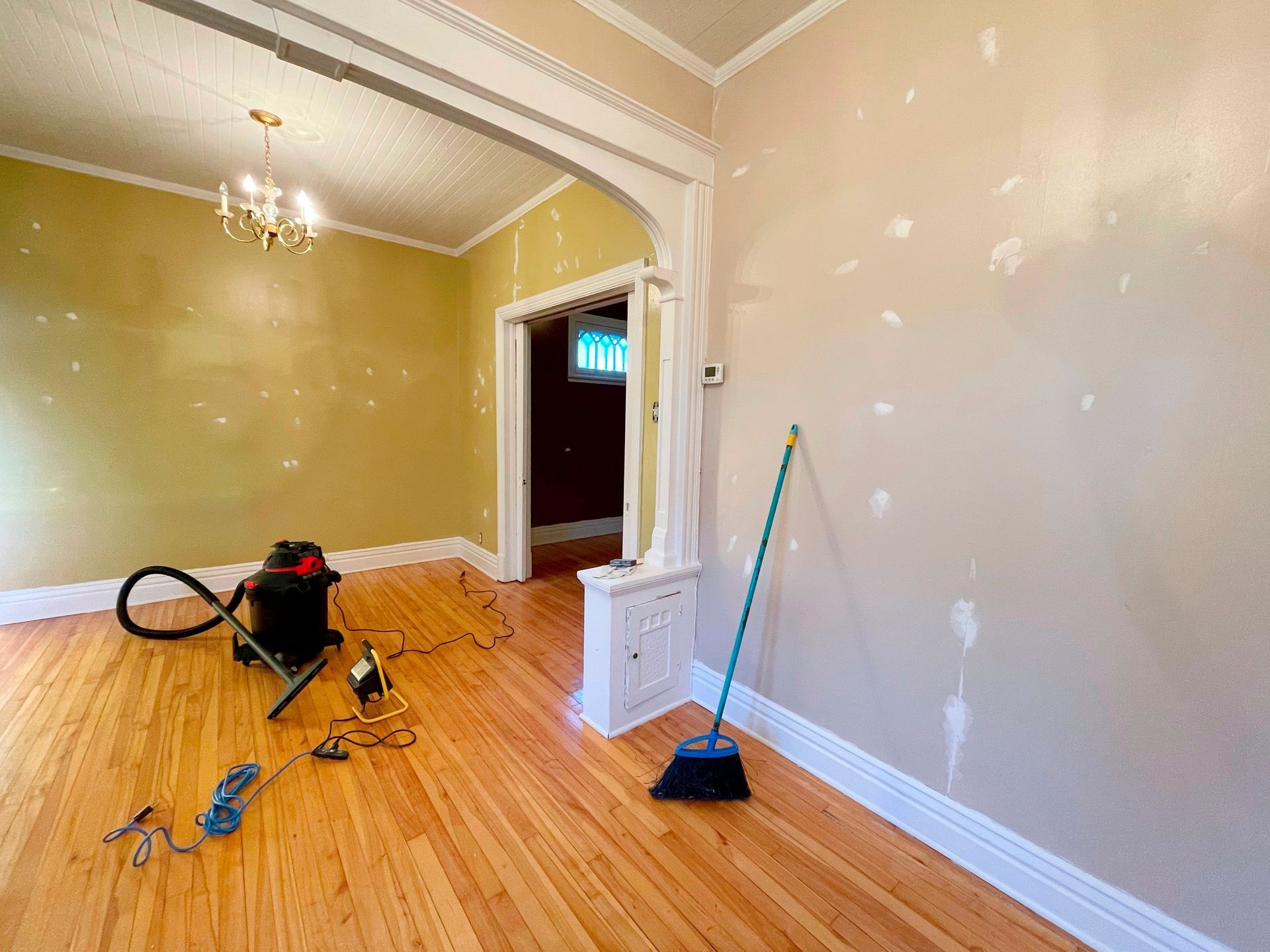 Room with hardwood floor, primed walls, and tools. Arched doorway, chandelier, broom and vacuum visible.