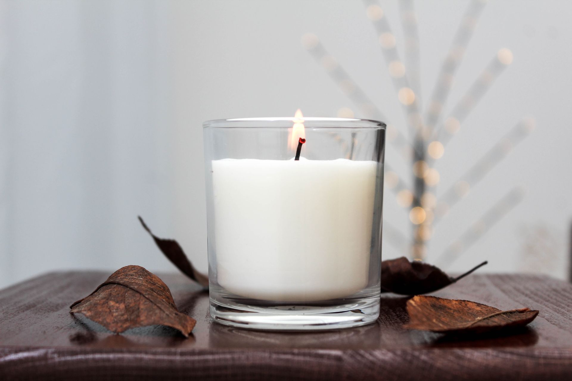 White candle in a glass jar on a brown surface with dried leaves, lit against a soft gray background