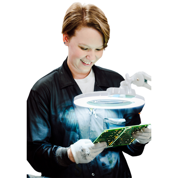 Woman examines circuit boards with a magnifying glass, smiling. She wears gloves and a dark jacket.