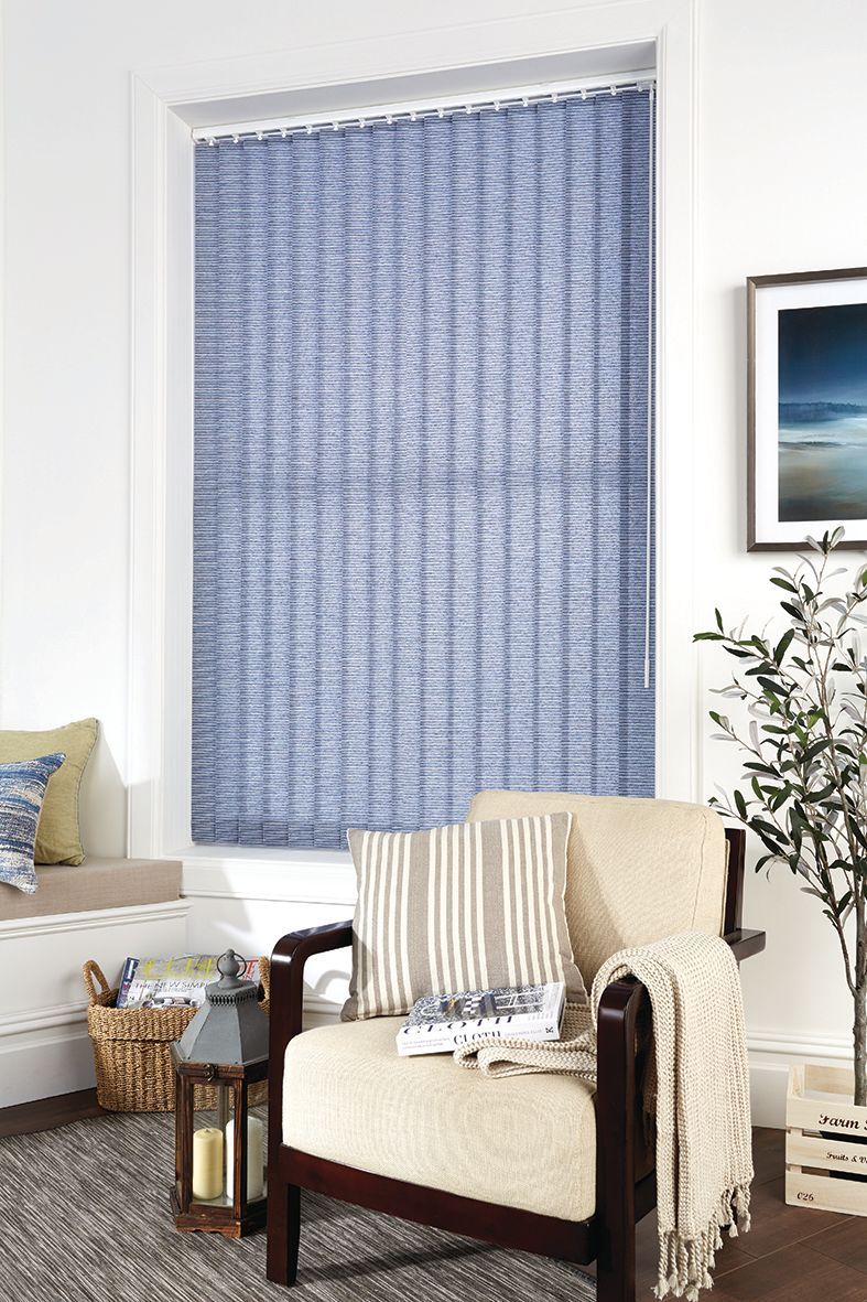 Blue woven vertical blinds on a window frame, a beige chair with pillows, a basket, and decor.