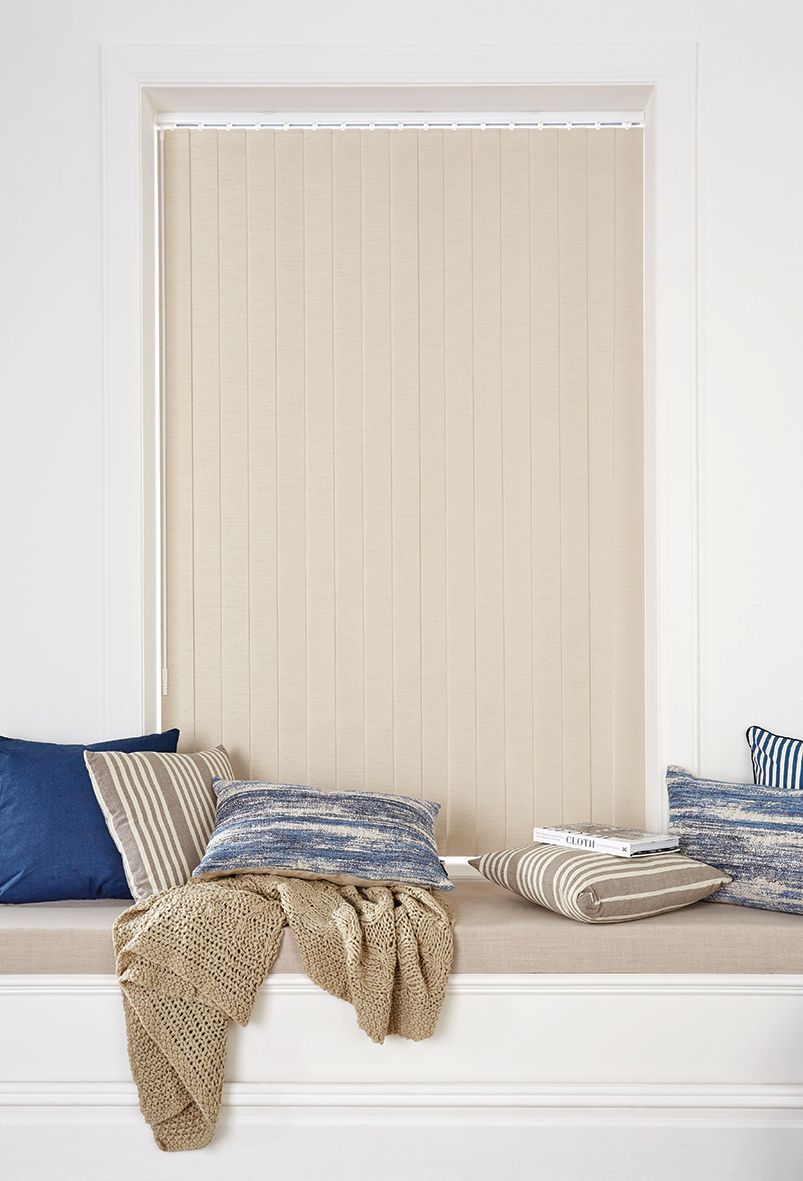 Beige vertical blinds in a white window frame, over a window seat with blue and patterned pillows, and a tan throw.