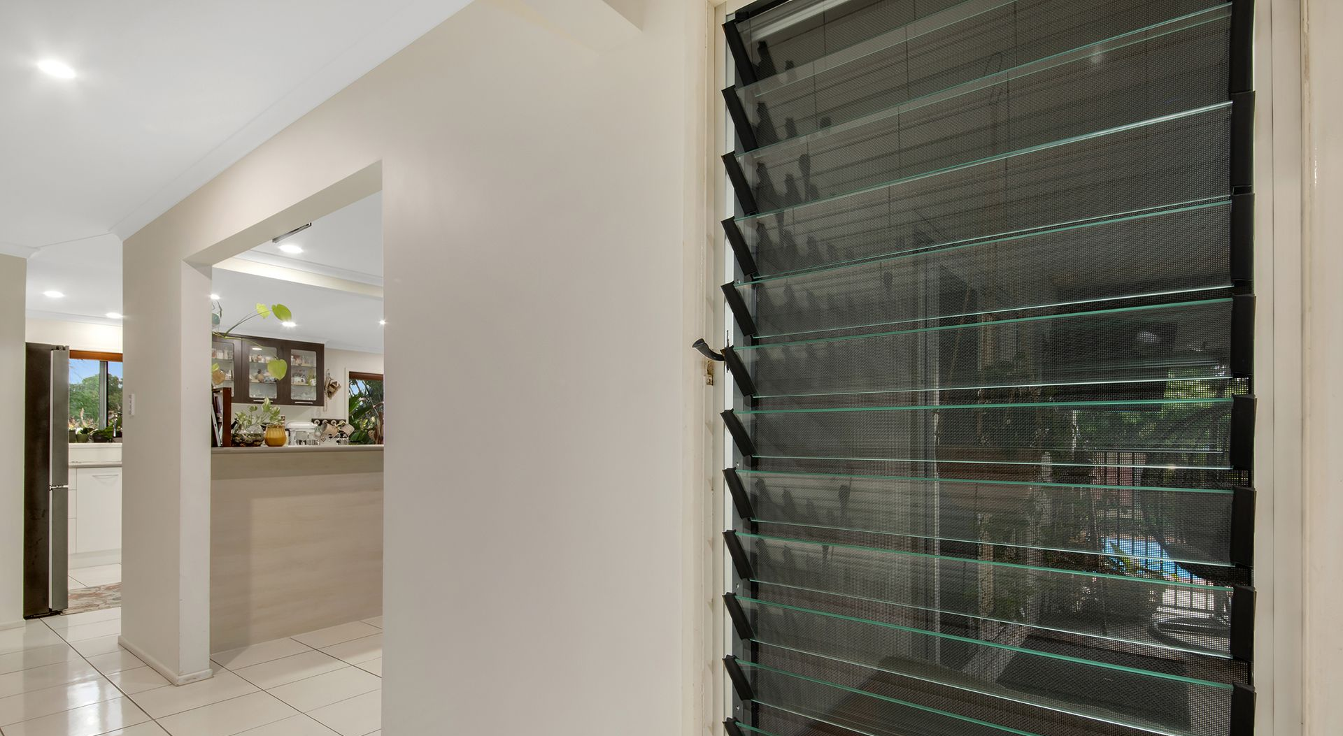 Hallway with a louvred window and view of a kitchen. White walls and tiled floor.