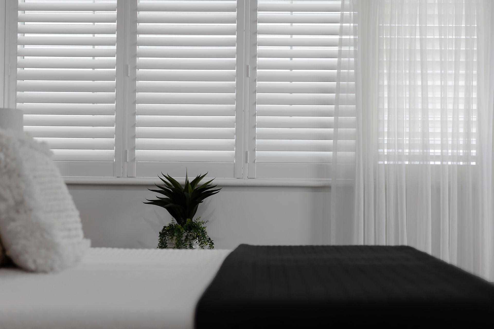 White shutters and sheer curtains over a window in a bedroom. A bed with a black throw and white pillows is in the foreground.