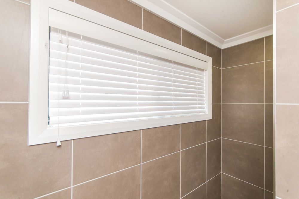 White Blinds in A Small Window in A Bathroom with Brown Tile Walls — Wares Blinds, Screens & Awnings In Gracemere, QLD
