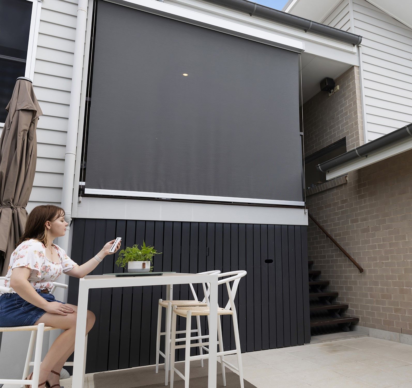 Woman operates a remote control, lowering a large black outdoor shade; sitting at a table Wares Blinds, Screens & Awnings In Gladstone Central, QLD