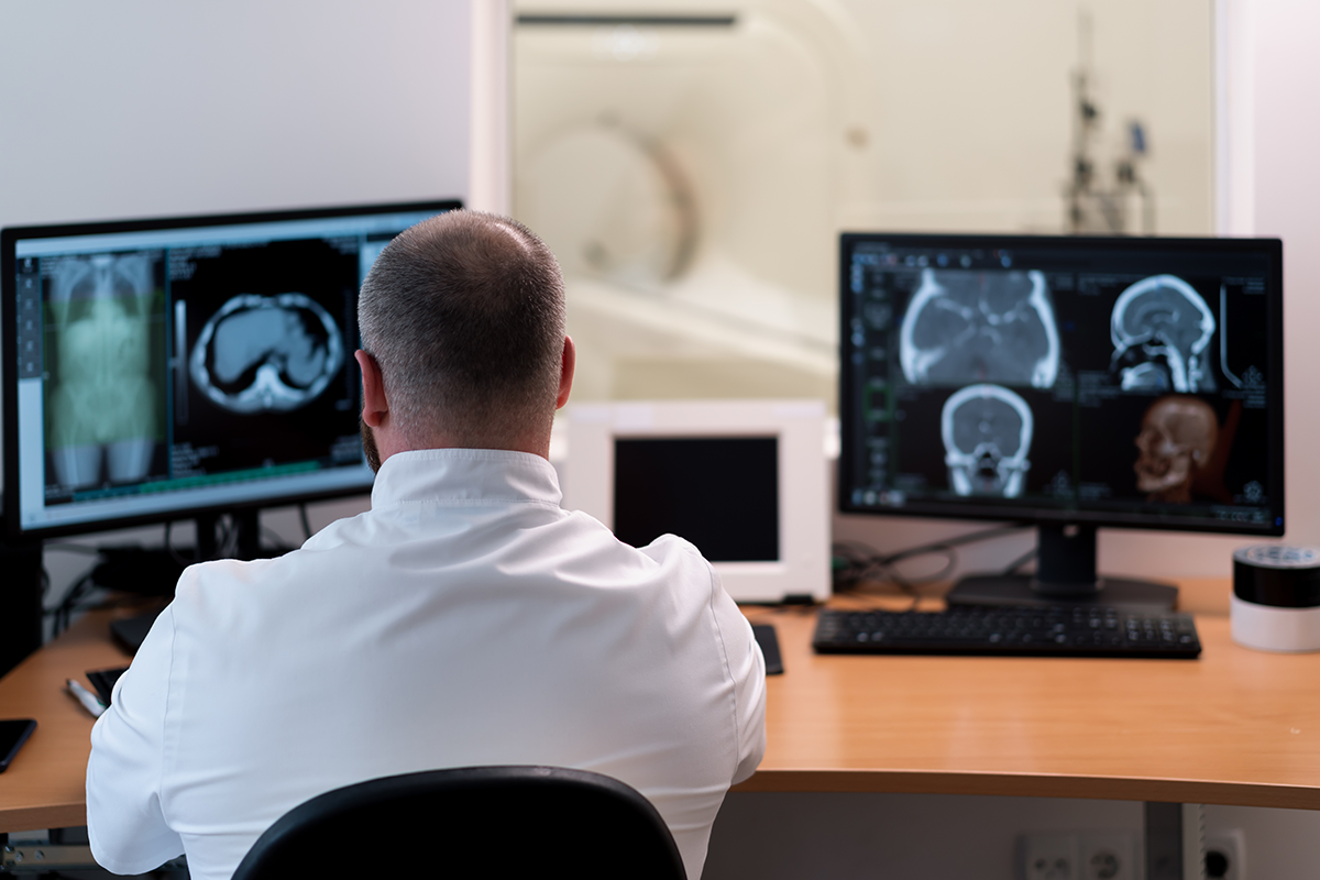 Radiologist reviewing CT scans of the chest and head on two monitors, with a CT scanner visible thro
