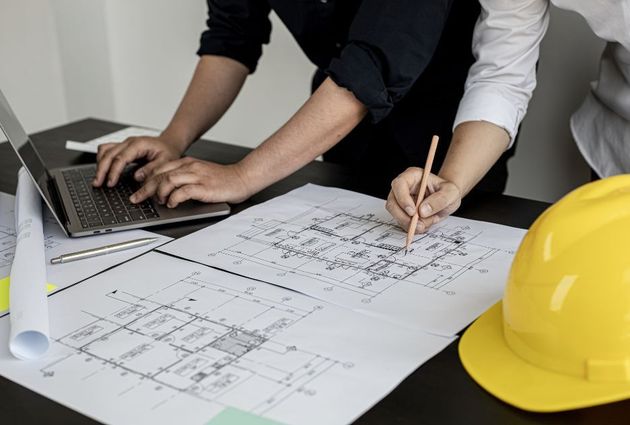 A man is using a laptop and a woman is writing on a blueprint.