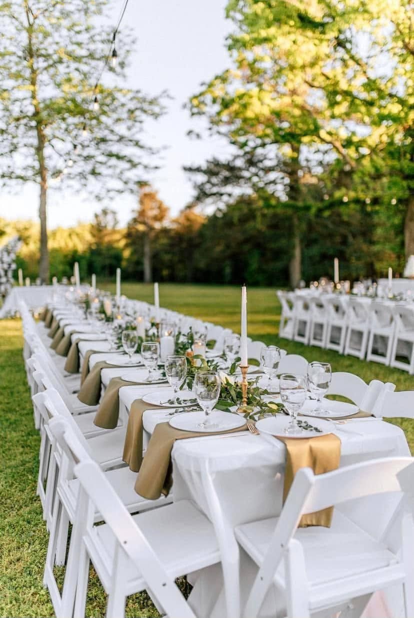 Outdoor wedding reception with long white table, white chairs, greenery centerpieces, and gold napkins on a lawn