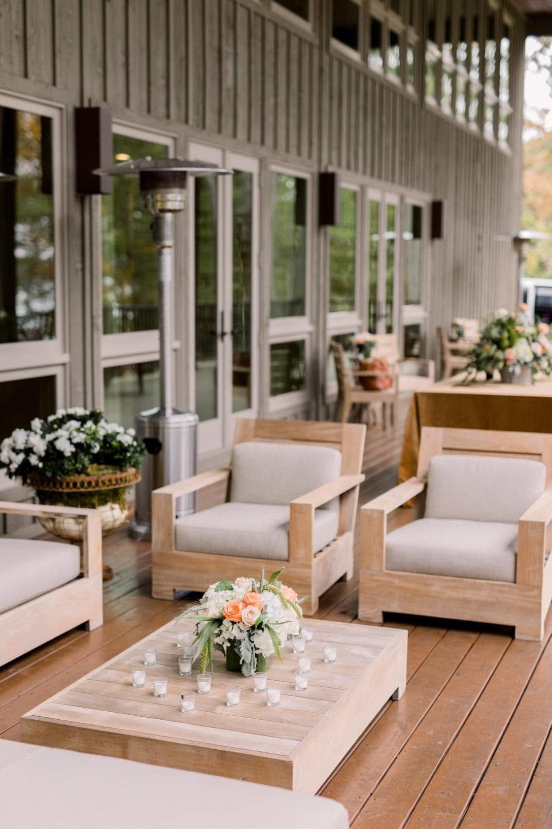 Outdoor patio with beige chairs, low coffee table, and floral arrangements on a wooden deck by a glass wall