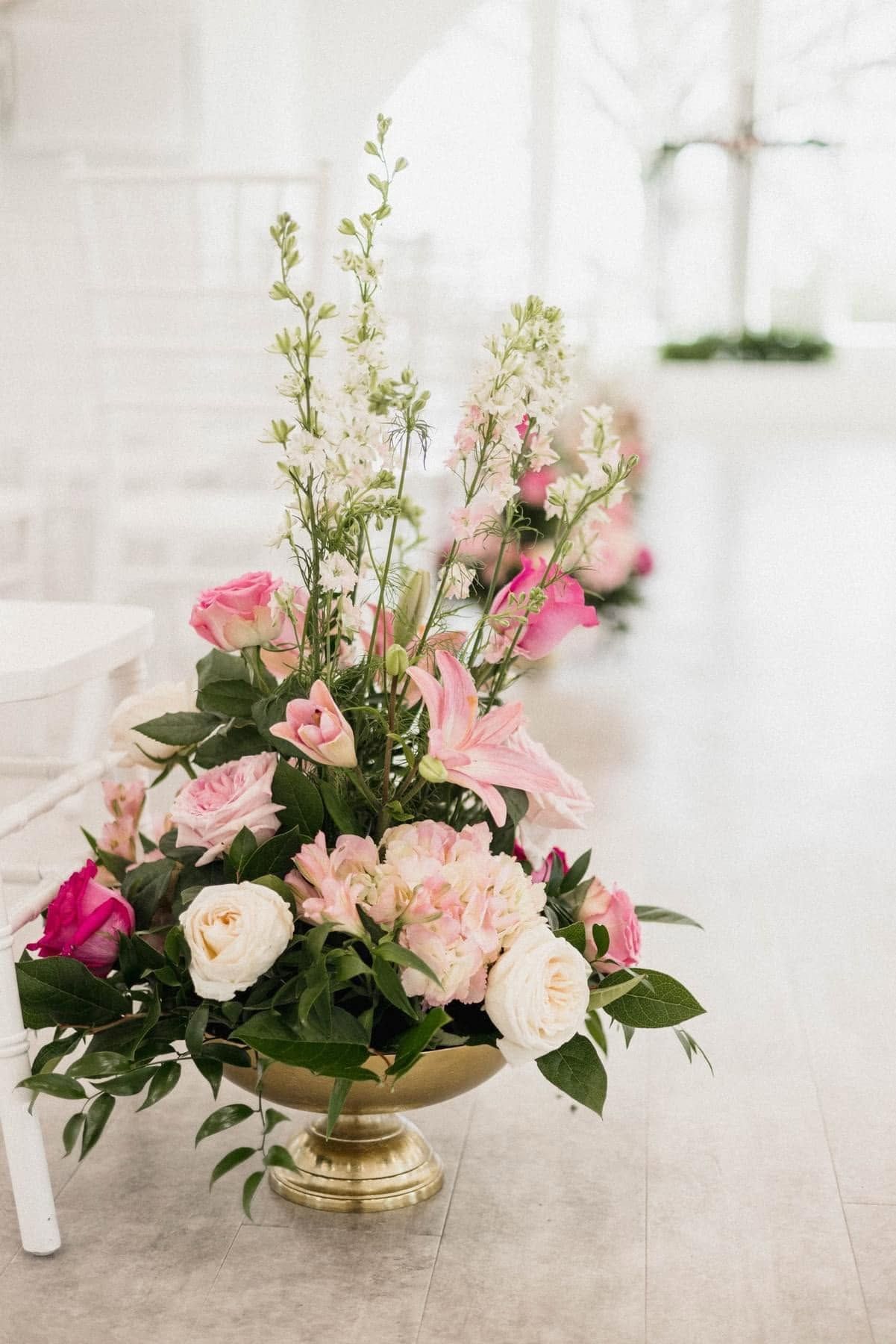 Pink and white floral arrangement in a gold vase on a white floor by a bright window