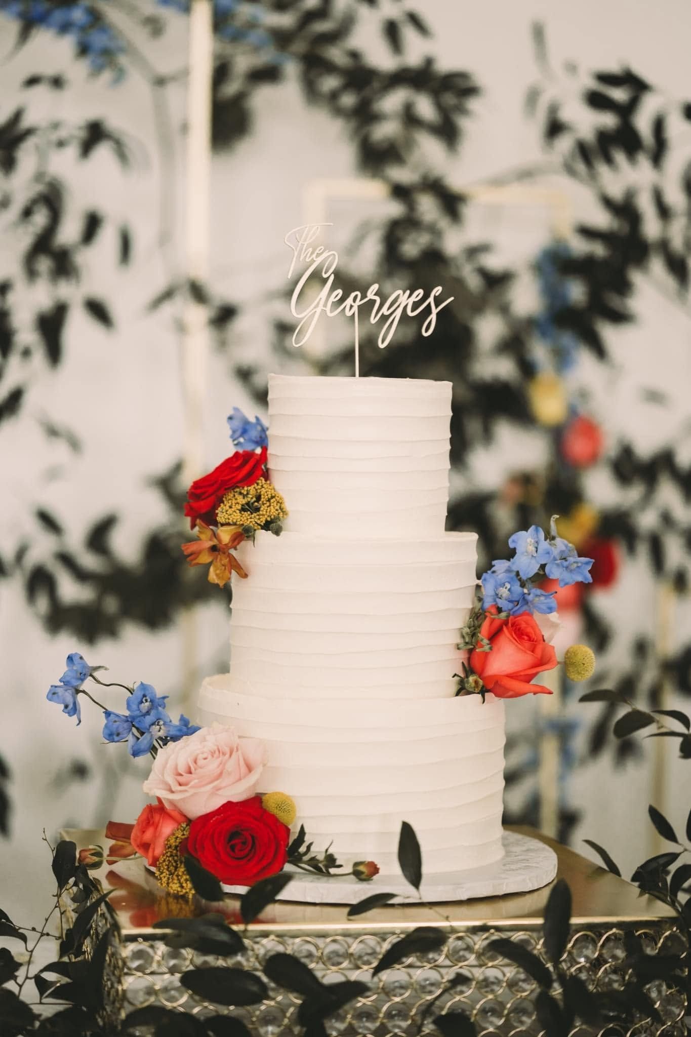 Tall white tiered cake with colorful flowers and a cursive topper on a decorative table