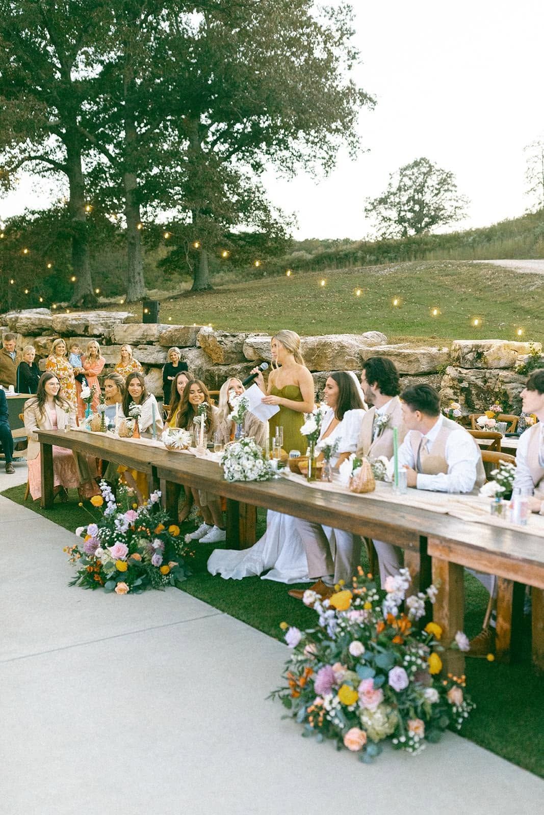Outdoor wedding reception with guests seated at a long wooden table decorated with flowers and candles