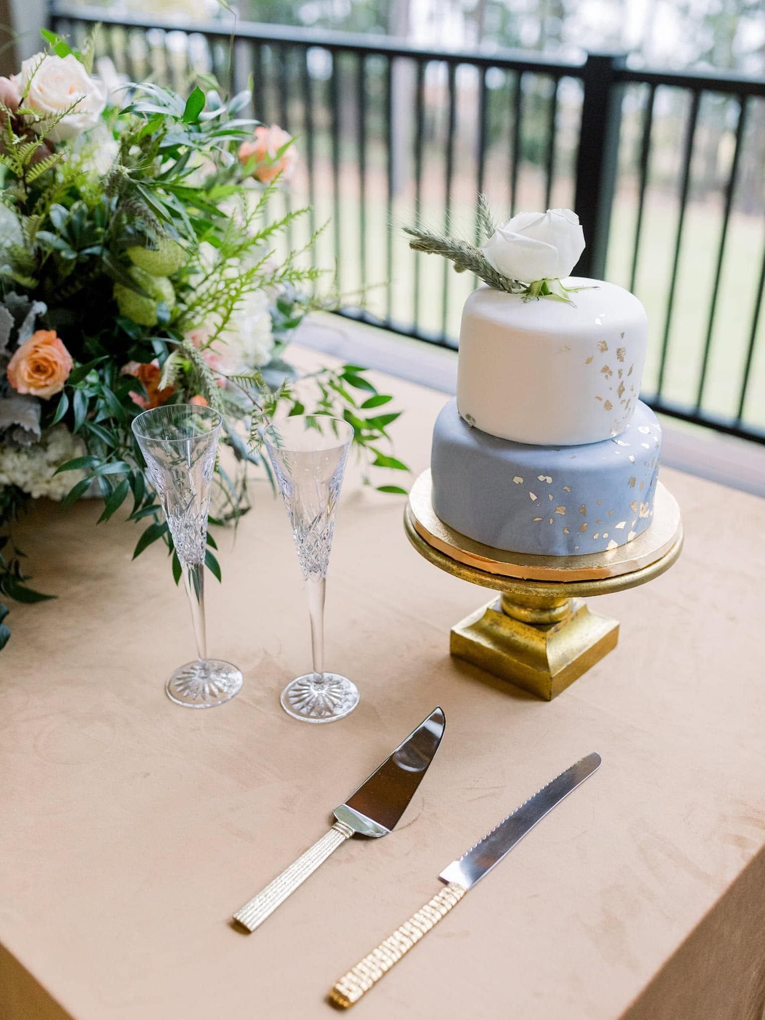 Two-tier white and blue cake with floral arrangement and serving tools on a decorated table outdoors