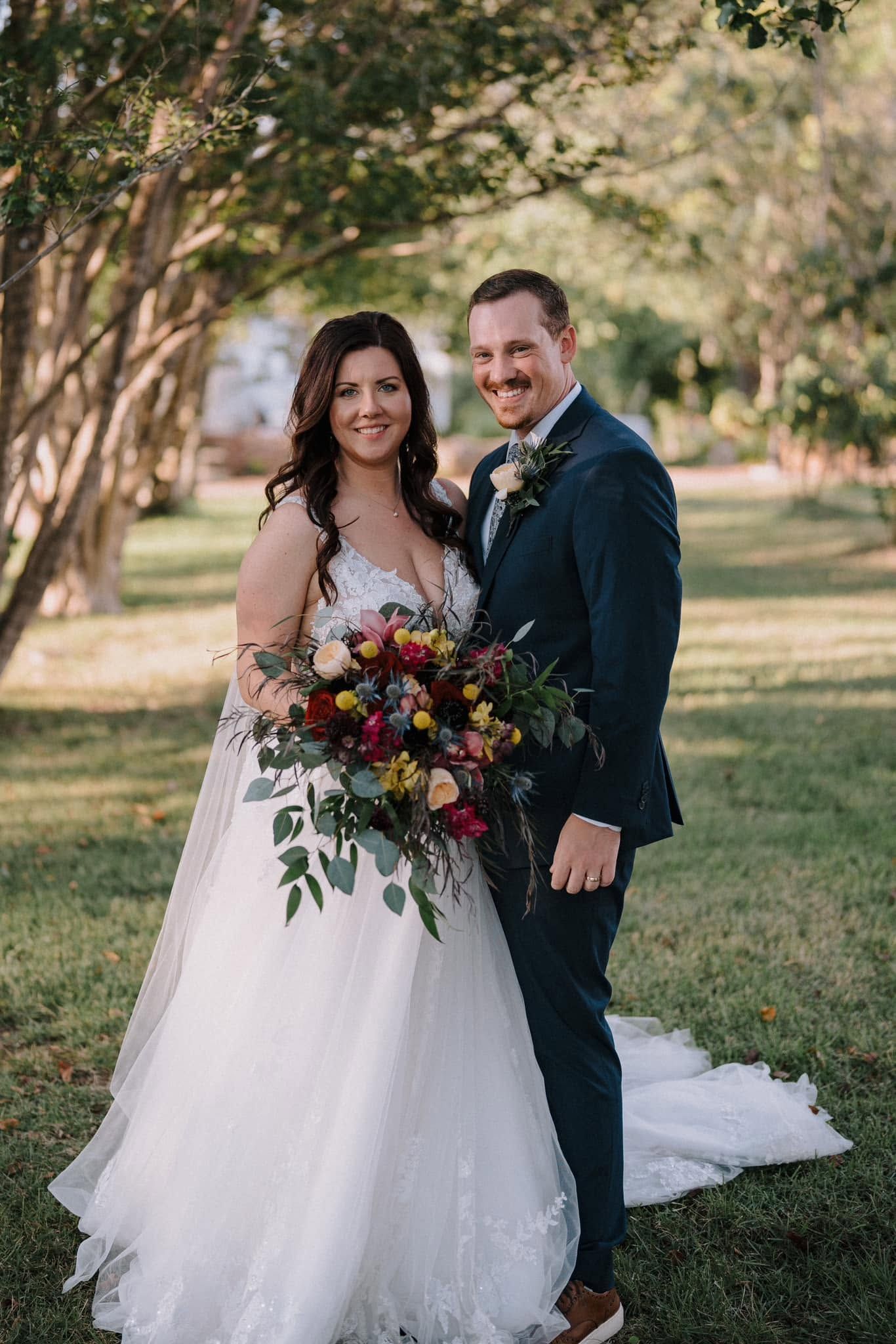 Bride and groom standing outdoors, smiling with a colorful bouquet and flowing white train