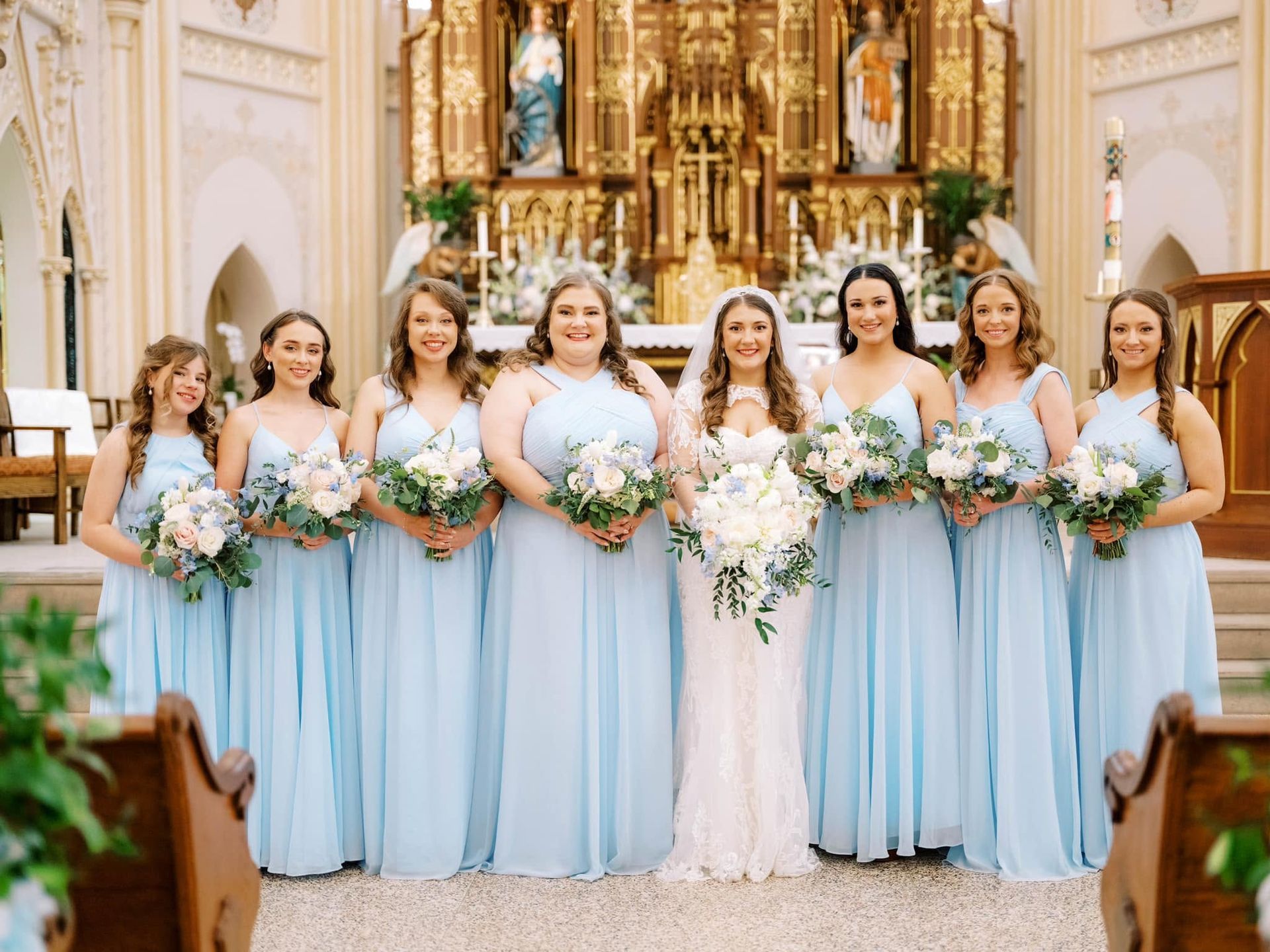 Bridesmaids in light blue dresses with bouquets stand with the bride in a white gown inside a church.
