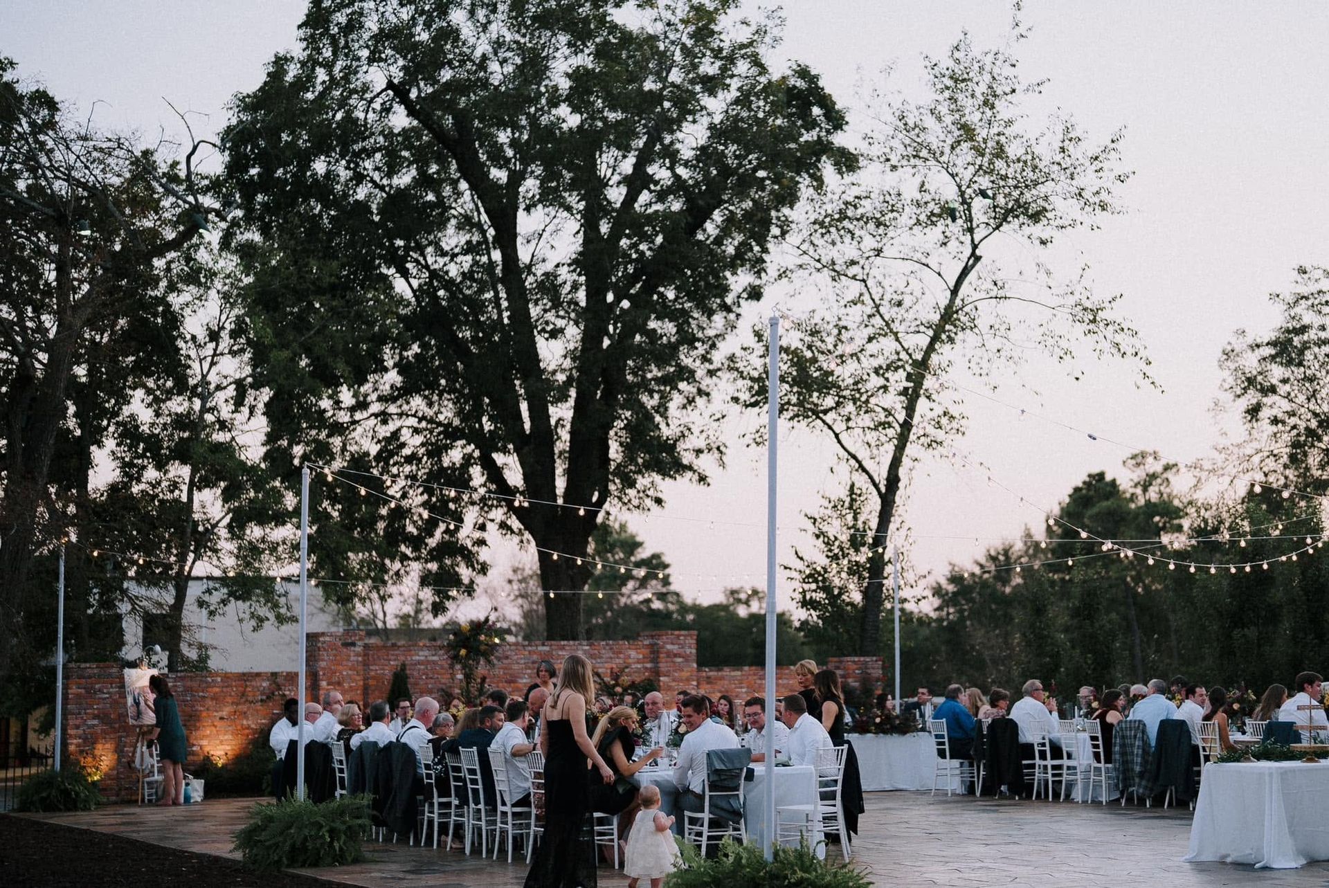 Outdoor evening reception with guests seated at round tables under large trees, lit by soft ambient lights.
