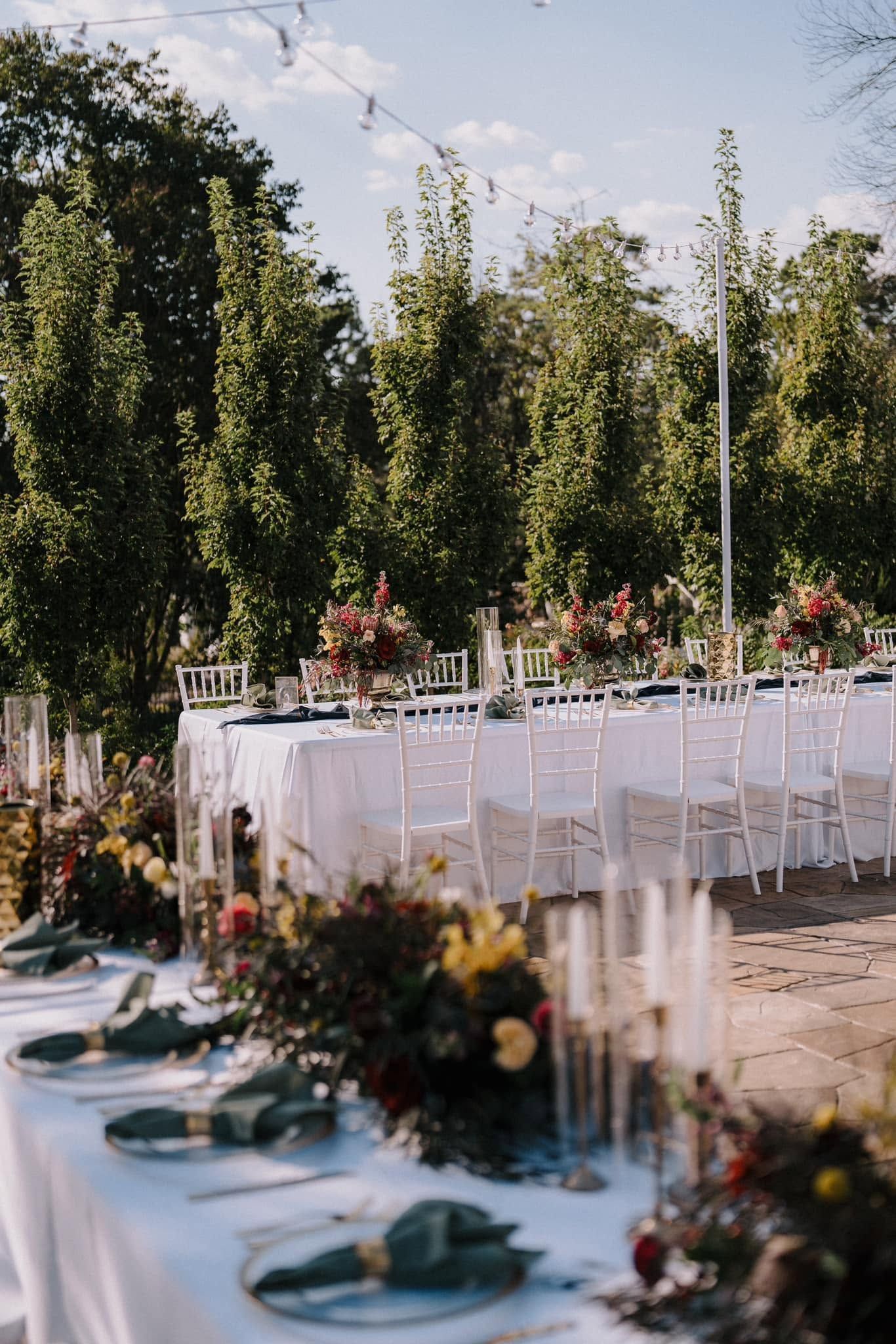 Outdoor banquet tables with white linens, glassware, flowers, and blue plates in a garden setting