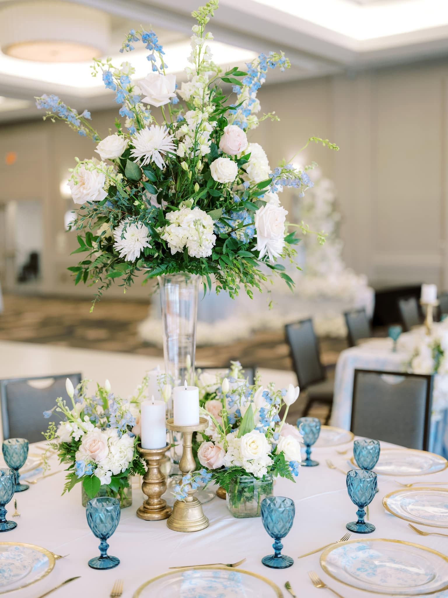 Elegant banquet table with white and blue floral centerpiece, gold accents, and blue glassware in a reception hall