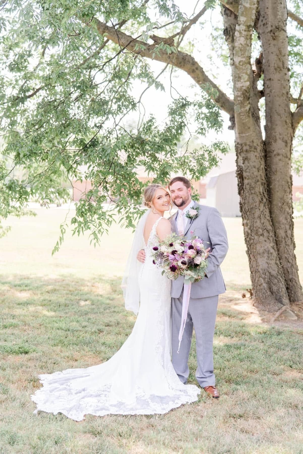 Bride and groom standing under a tree, holding a bouquet on a sunny lawn.