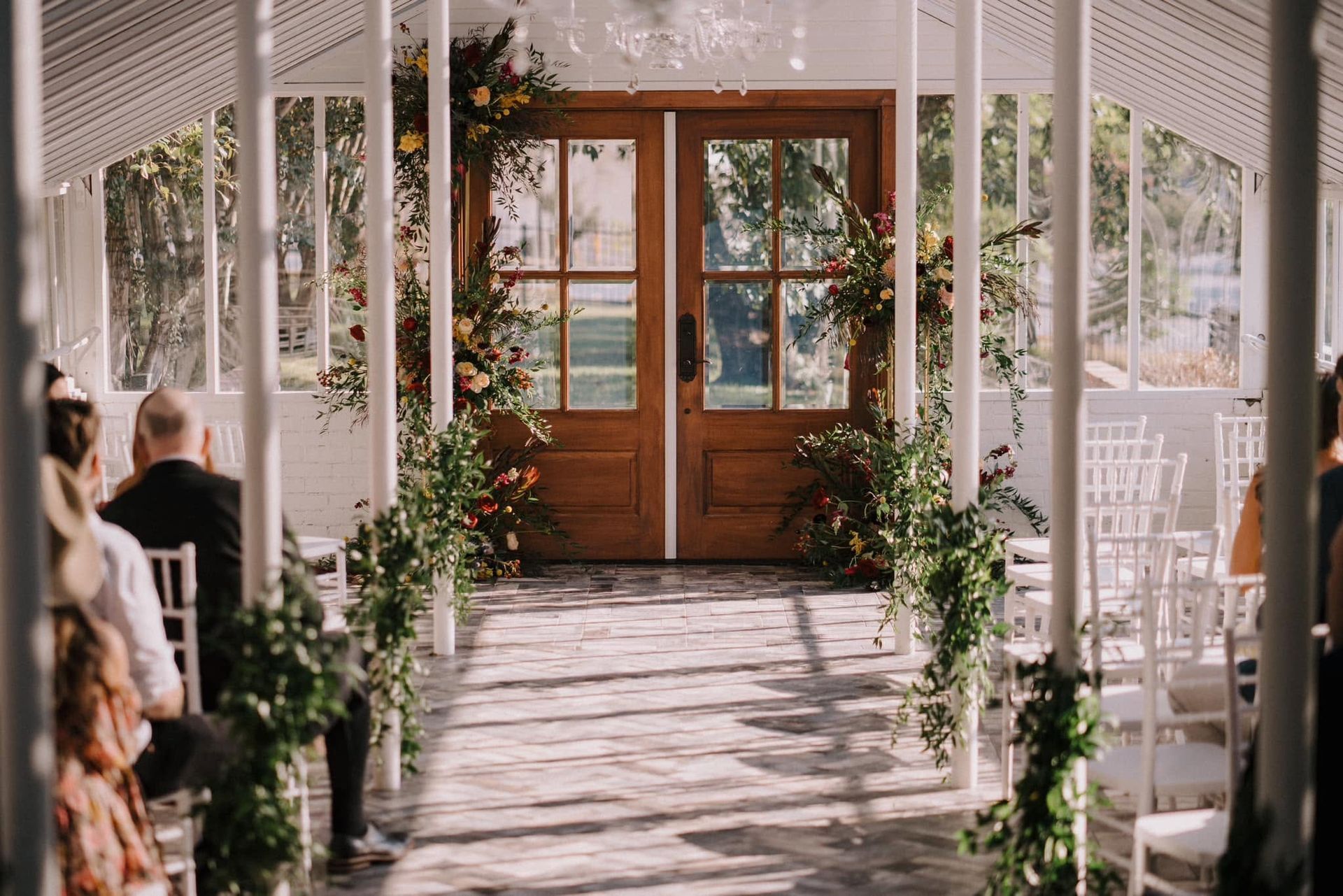Sunlit wedding aisle in a white garden pavilion with wooden doors and floral decor