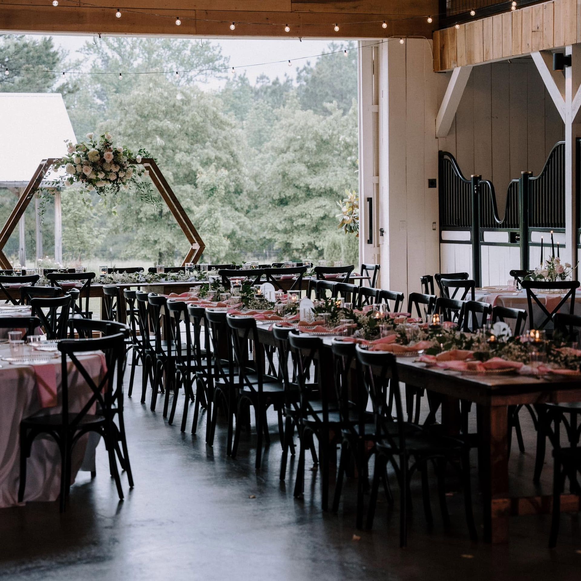 Decorated indoor event venue with long banquet tables, black chairs, and floral arch by large windows