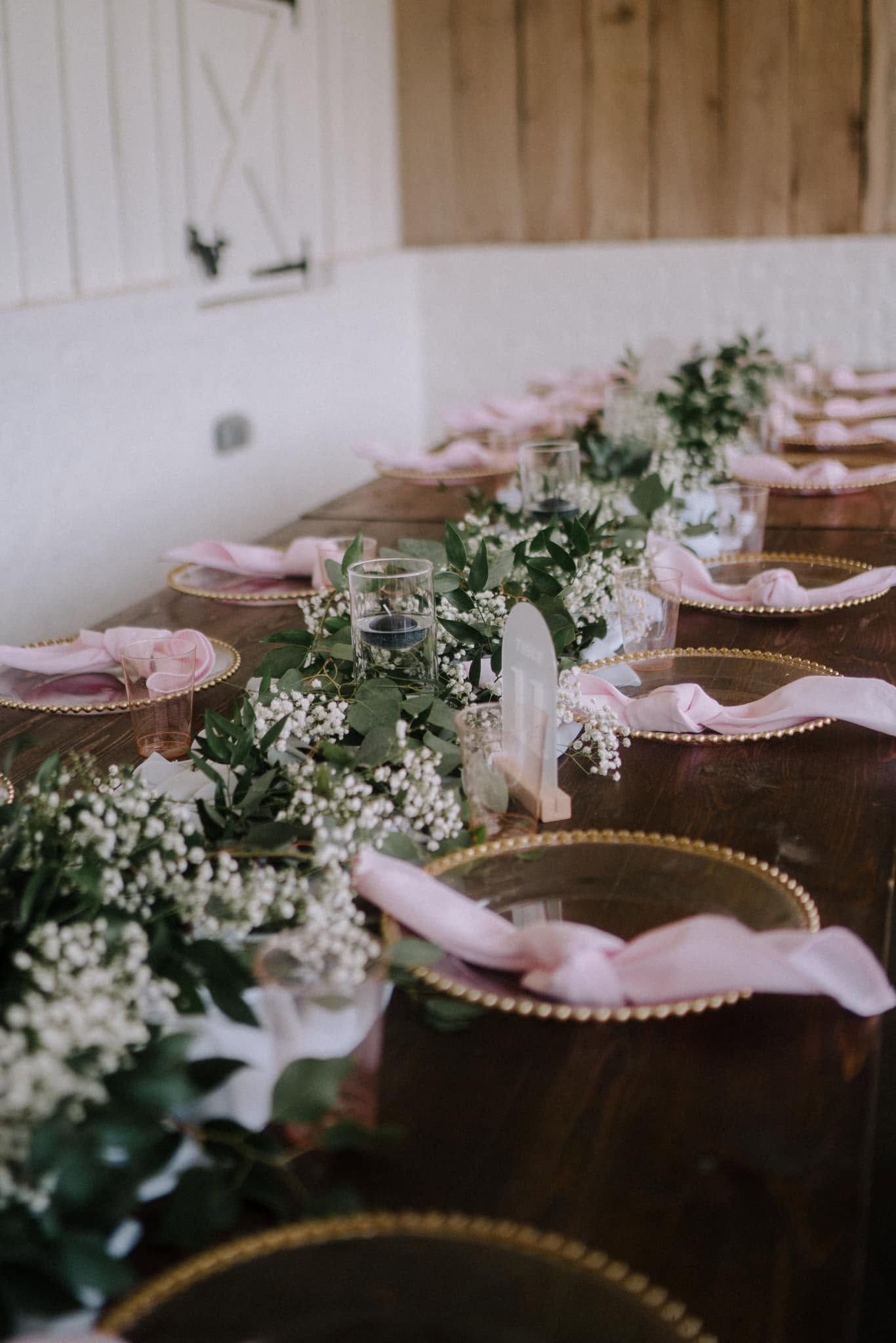 Long dining table set with pink napkins, white floral garlands, and place settings in a rustic room