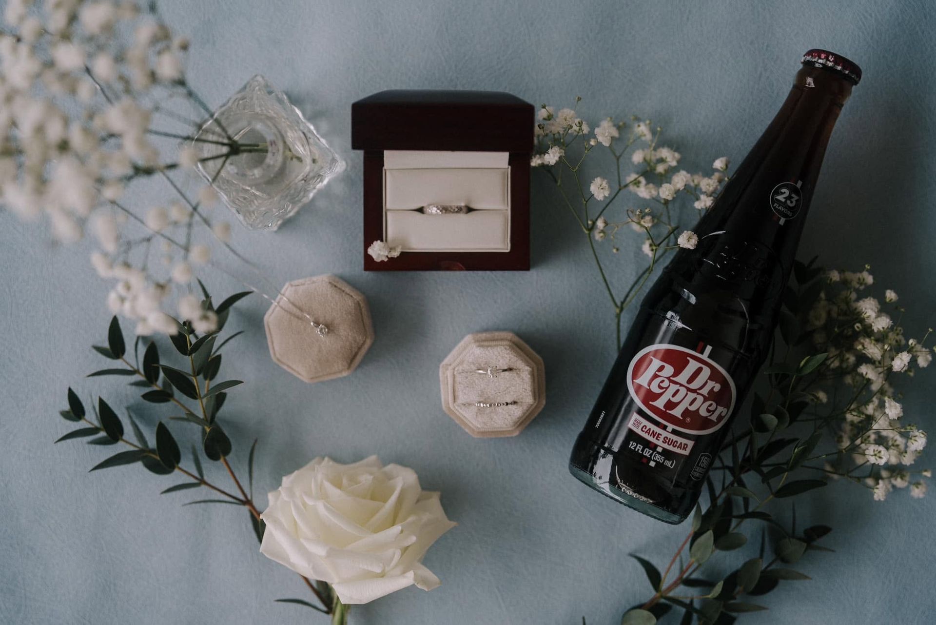 Flat lay of wedding accessories: rings, floral accents, and a champagne bottle on a blue background