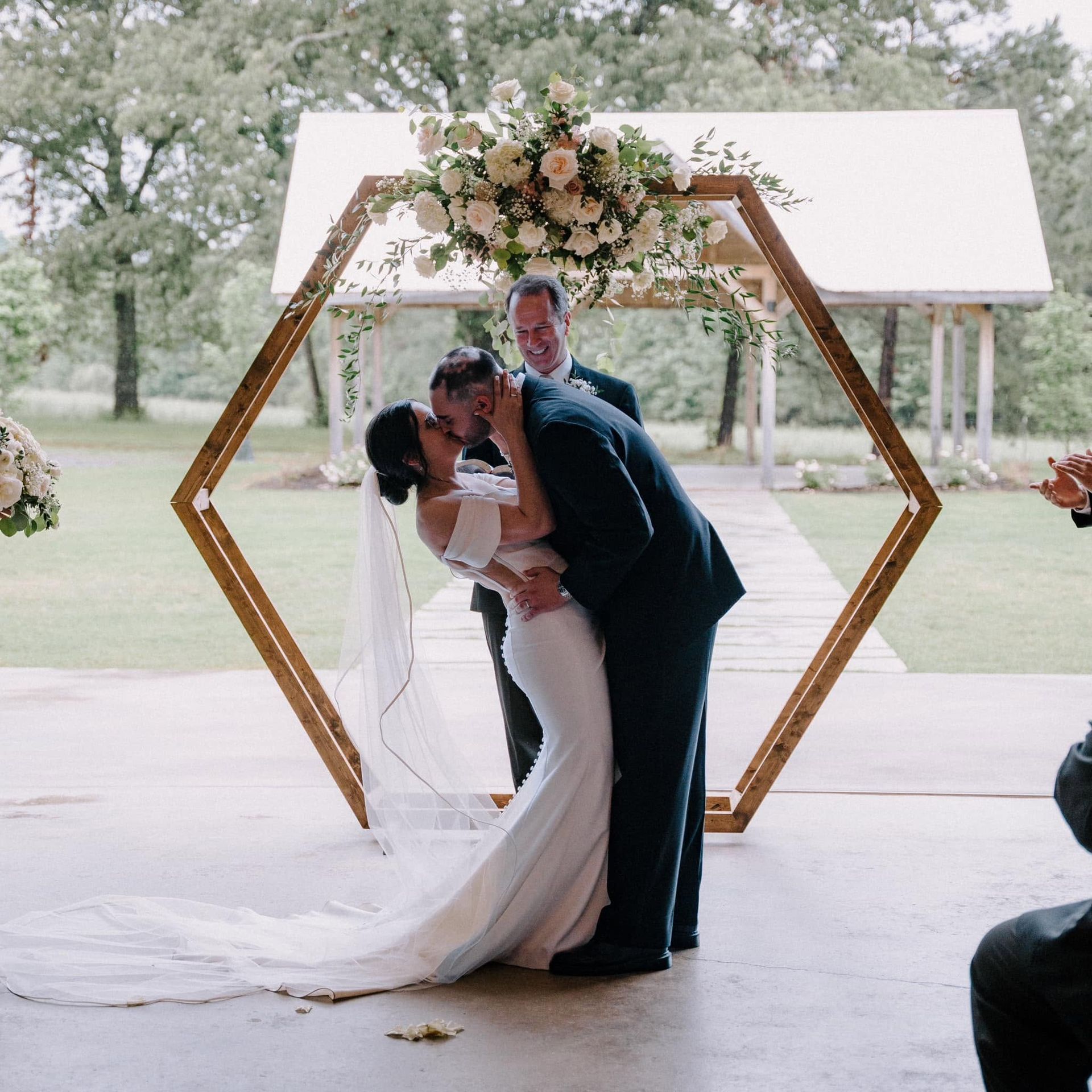 Bride and groom kiss beneath a floral hexagon arch at an outdoor wedding ceremony