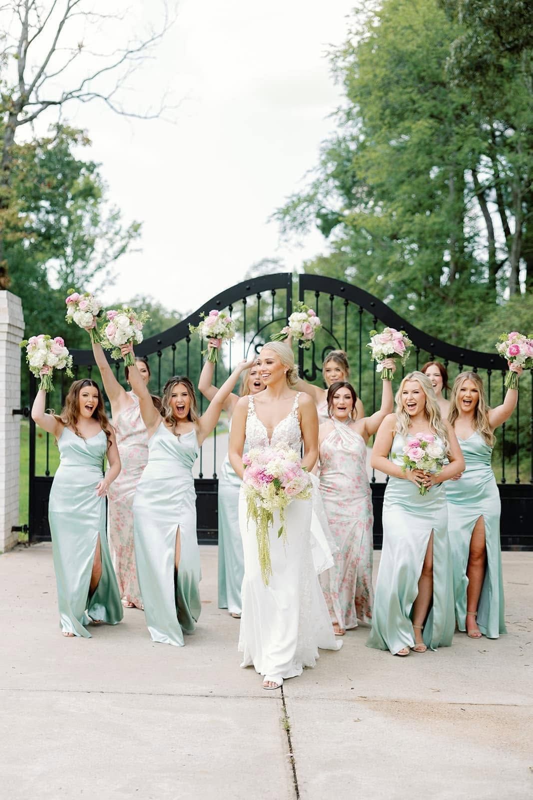 Bridal party in white and sage dresses poses outdoors in front of a black gate.
