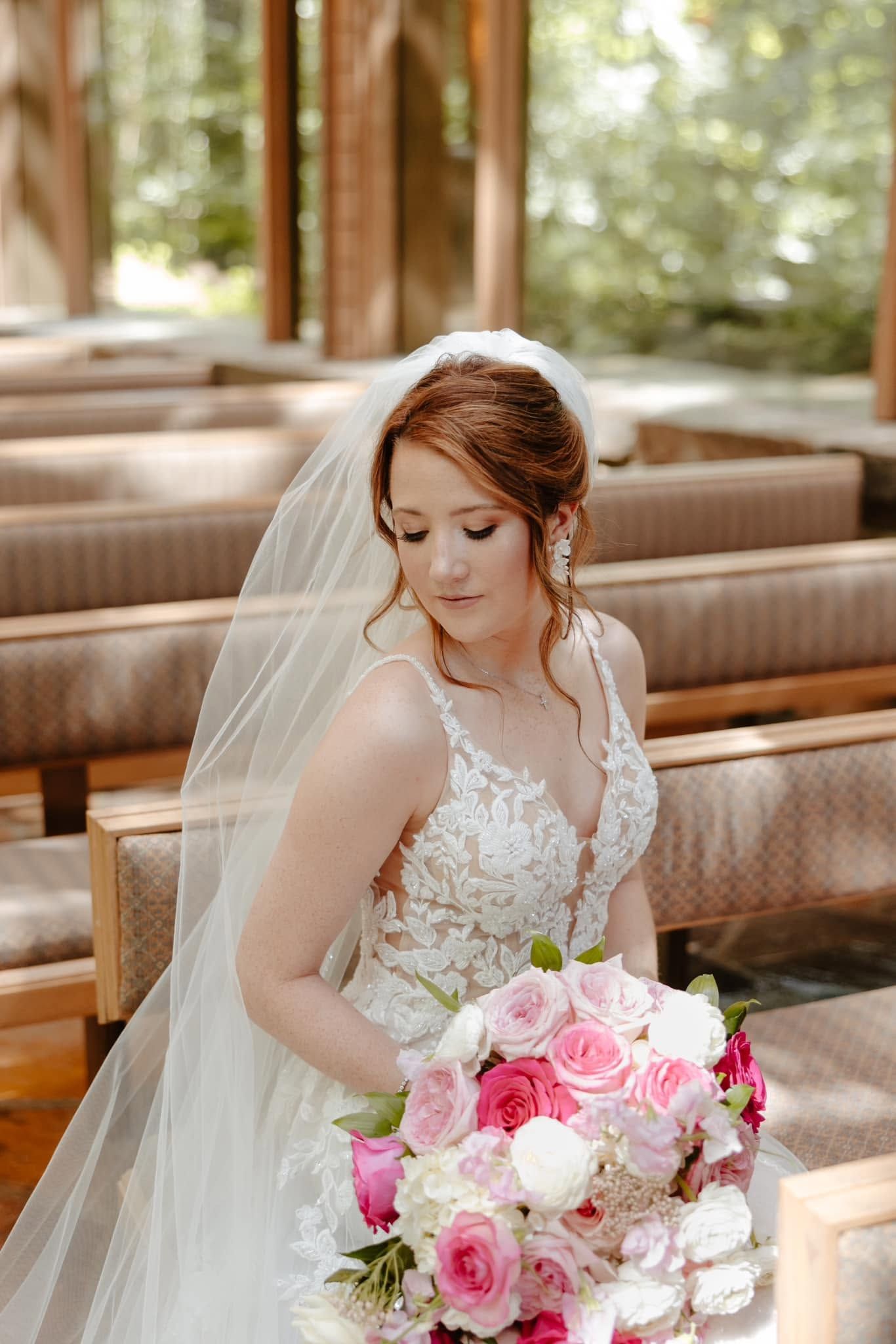 Bride in a white lace gown holding a pink and white bouquet, seated on a wooden bench outdoors.