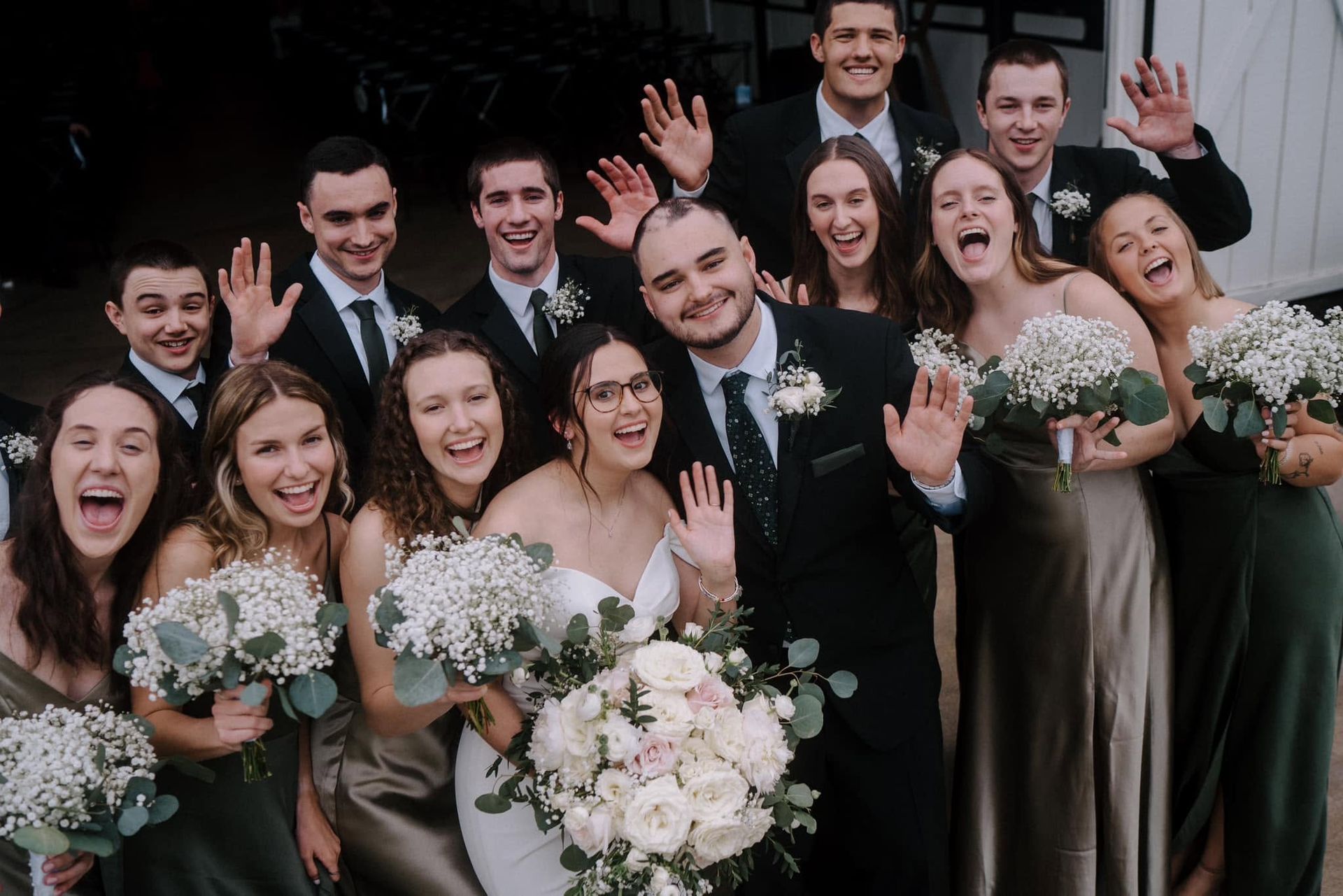 Wedding party smiling and cheering, holding white bouquets in front of a dark backdrop.