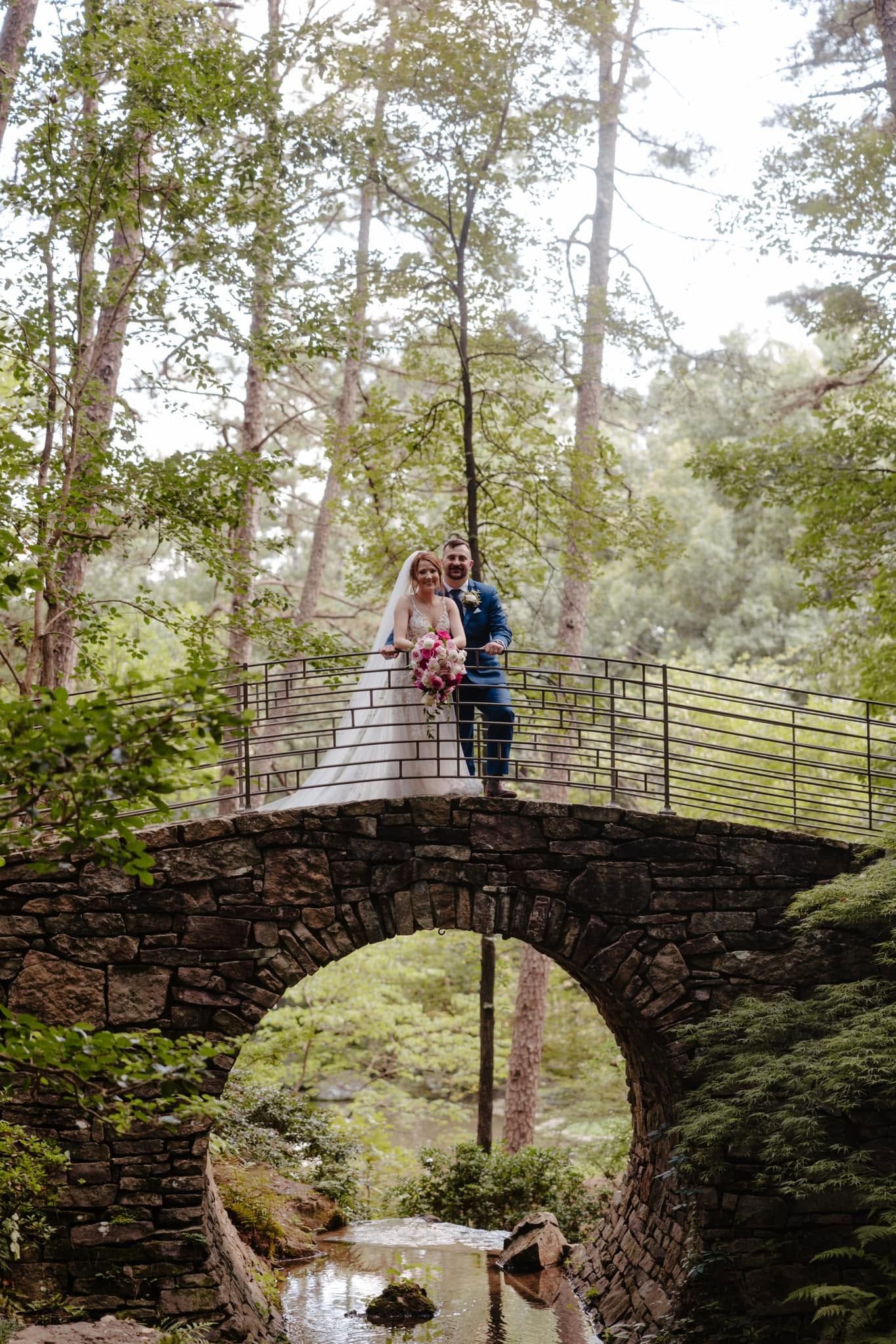 Two people standing on a stone bridge in a wooded garden, with a stream below.