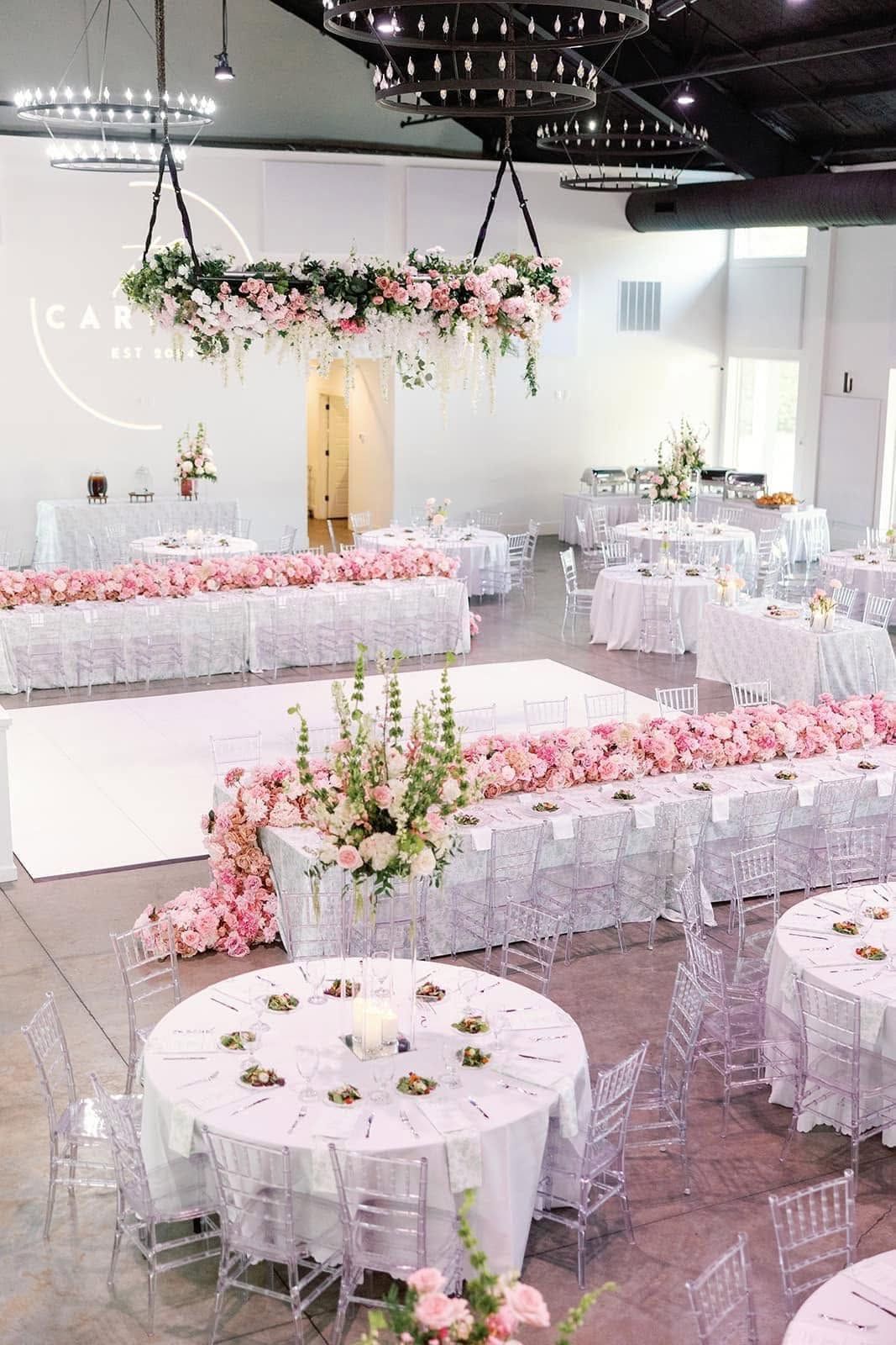 Elegant indoor wedding reception with white tables, pink floral decor, and sheer white chairs
